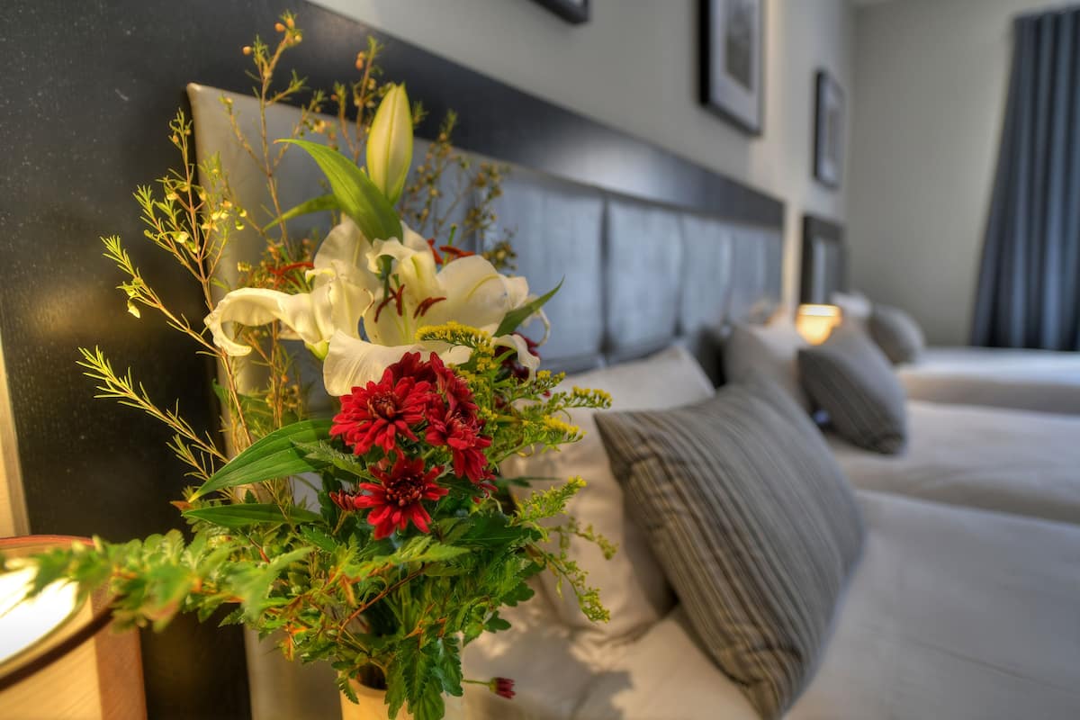 A hotel room with a bed, a bedside table with a lamp, and a decorative floral arrangement on the nightstand.