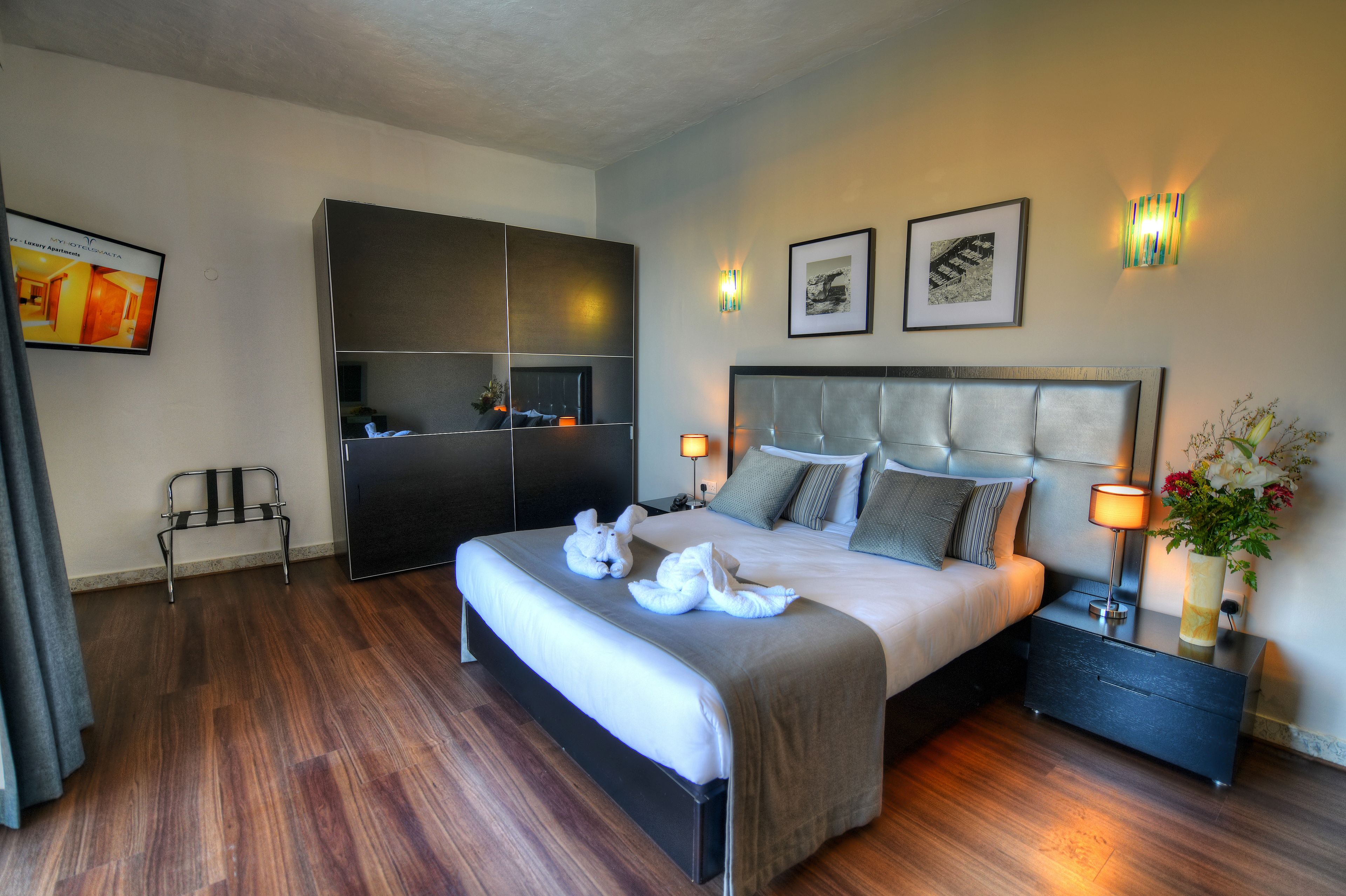A modern hotel room with a large bed, a bedside table with a lamp, a small seating area, and artwork on the walls.