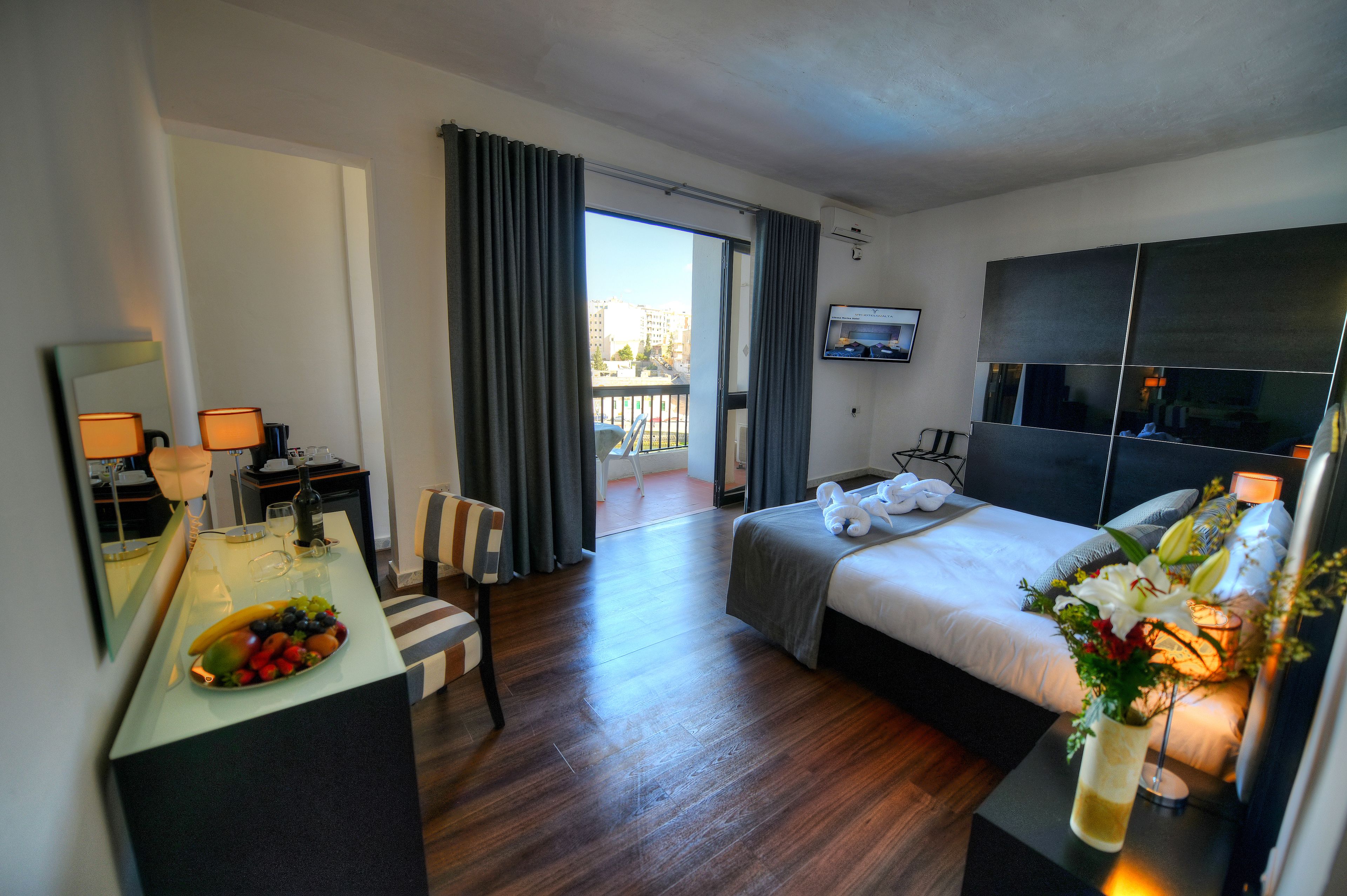 A hotel room with a bed, a desk with a fruit bowl, a chair, a TV, and a balcony with curtains.