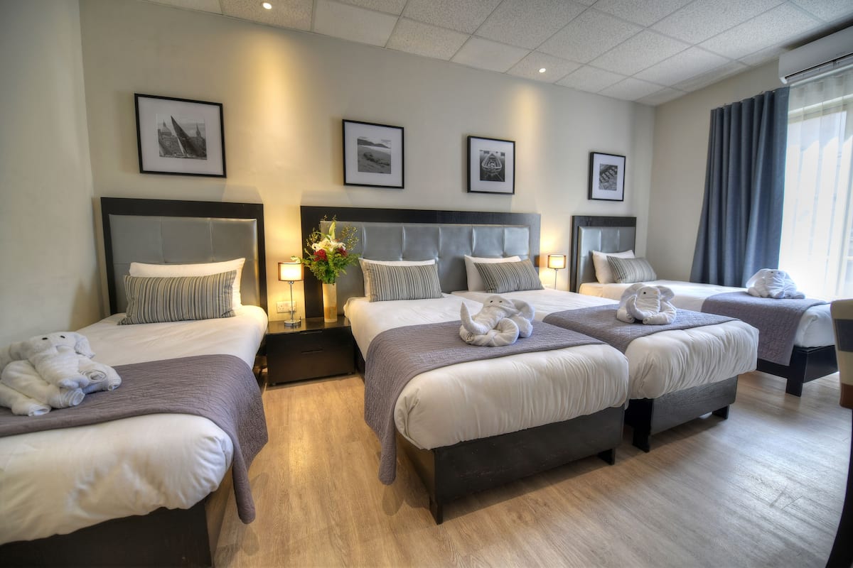 A hotel room with three beds, each with a headboard, a bedside table, and a lamp. There are framed pictures on the wall and a window with curtains.