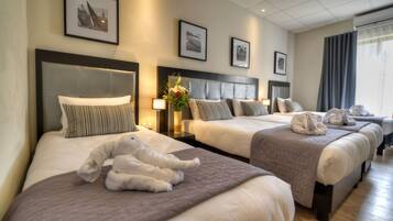 A hotel room with three beds, each with a headboard, bedside tables, and lamps. There are framed pictures on the wall and a window with curtains.