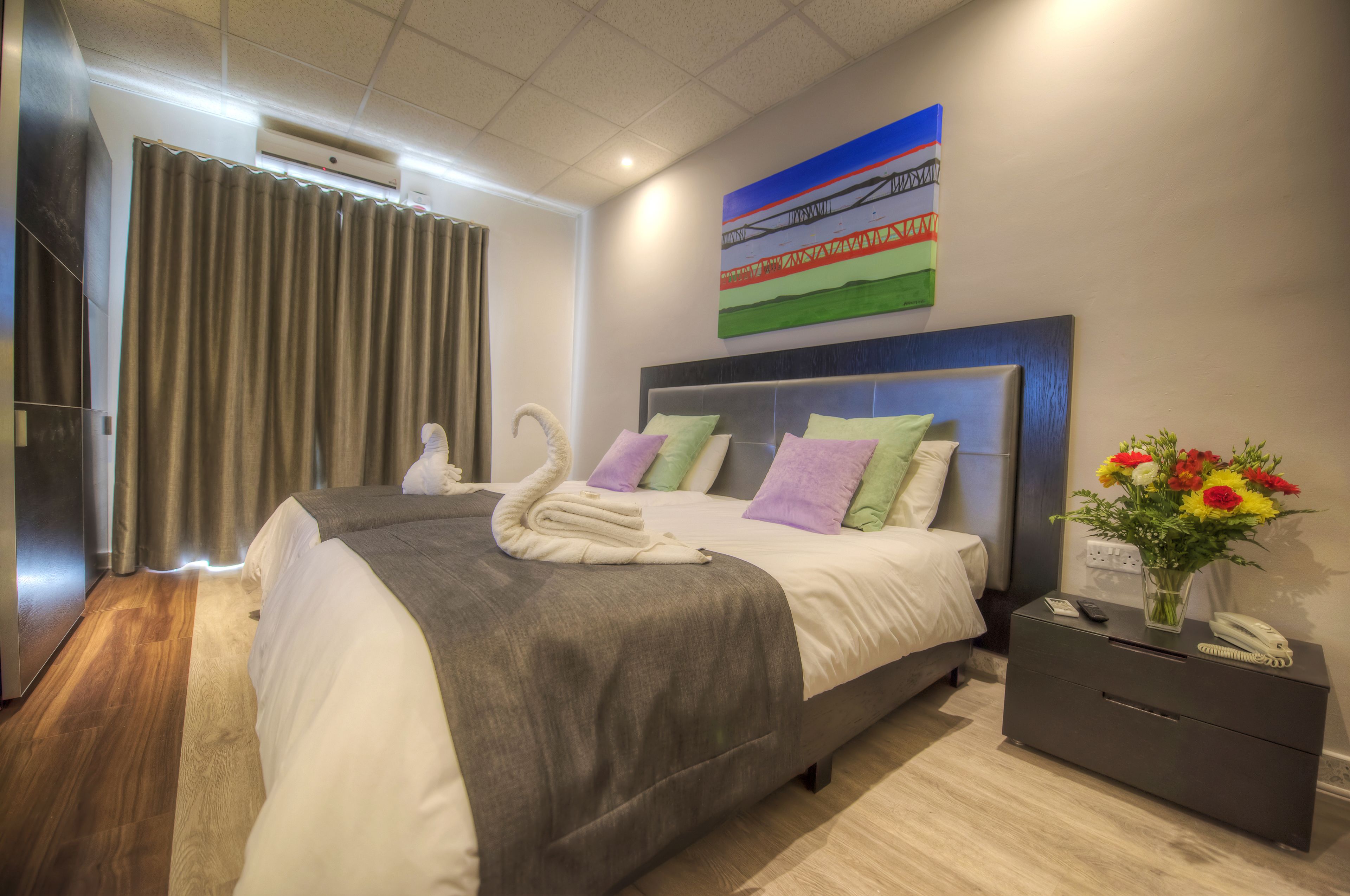 A modern hotel room with a large bed, a nightstand with a phone and vase of flowers, and a painting on the wall.
