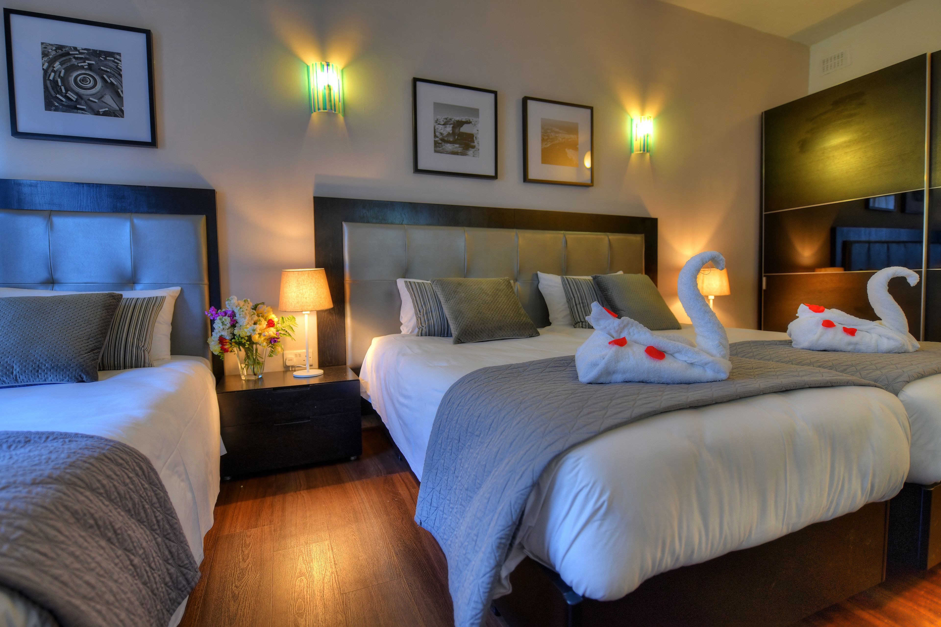 A hotel room with two beds, a nightstand with a lamp, framed pictures on the wall, and a large mirror.
