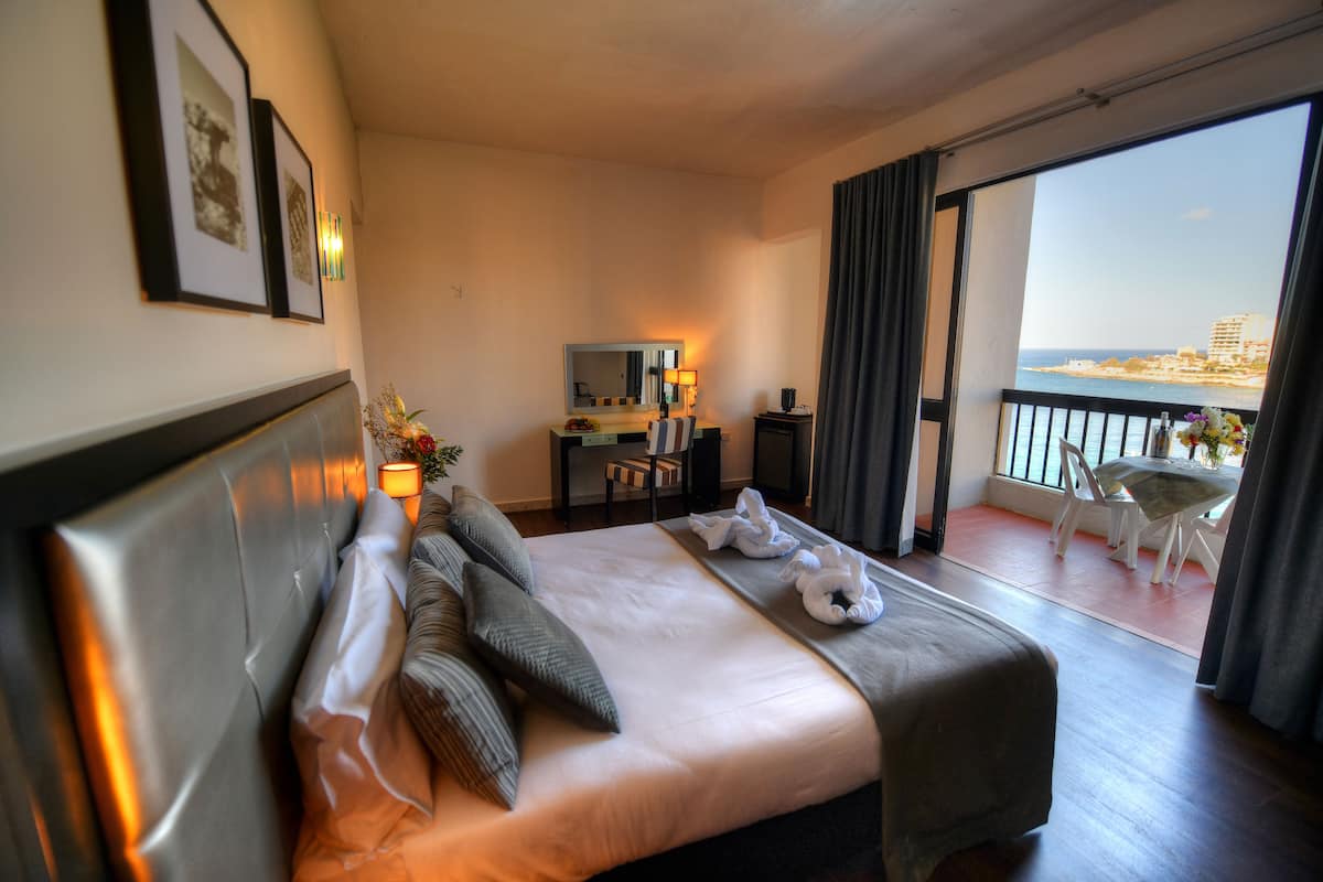 A hotel room with a bed, a desk with a lamp, a chair, and a view of the sea.