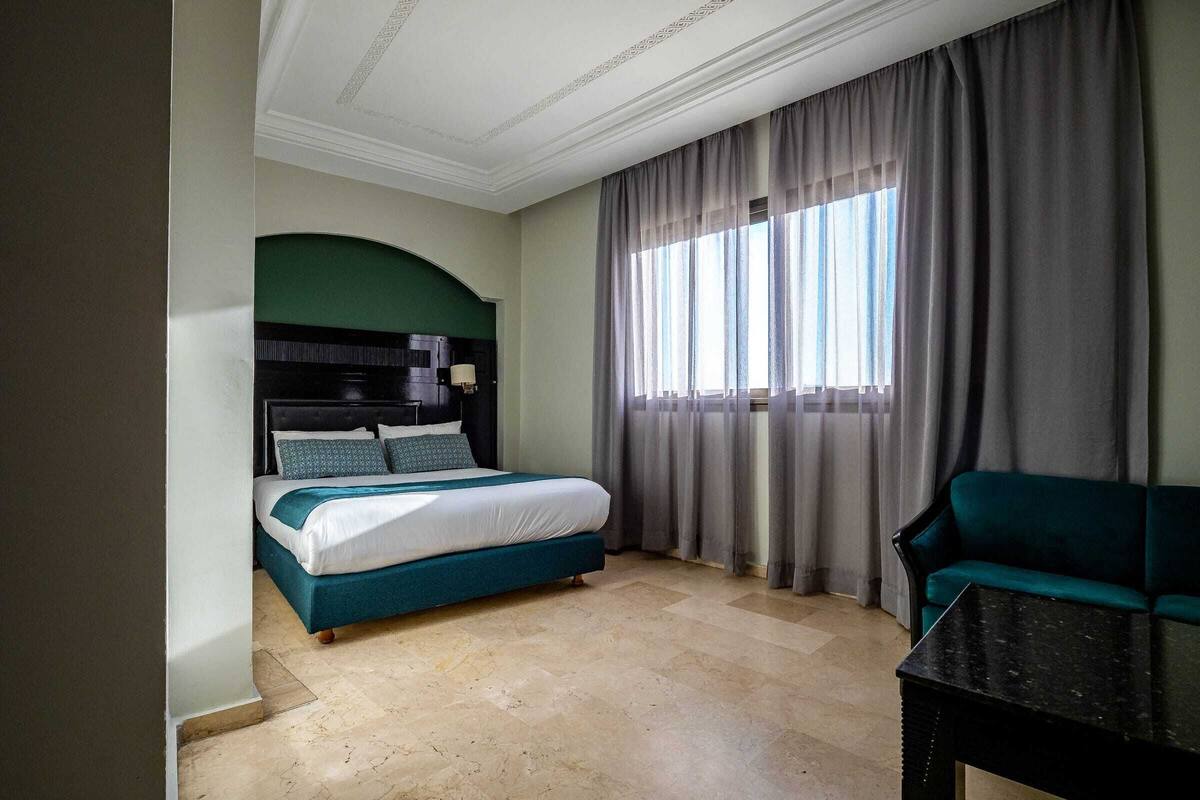 Une chambre d’hôtel avec un lit, un canapé vert, une table et une fenêtre avec des rideaux.