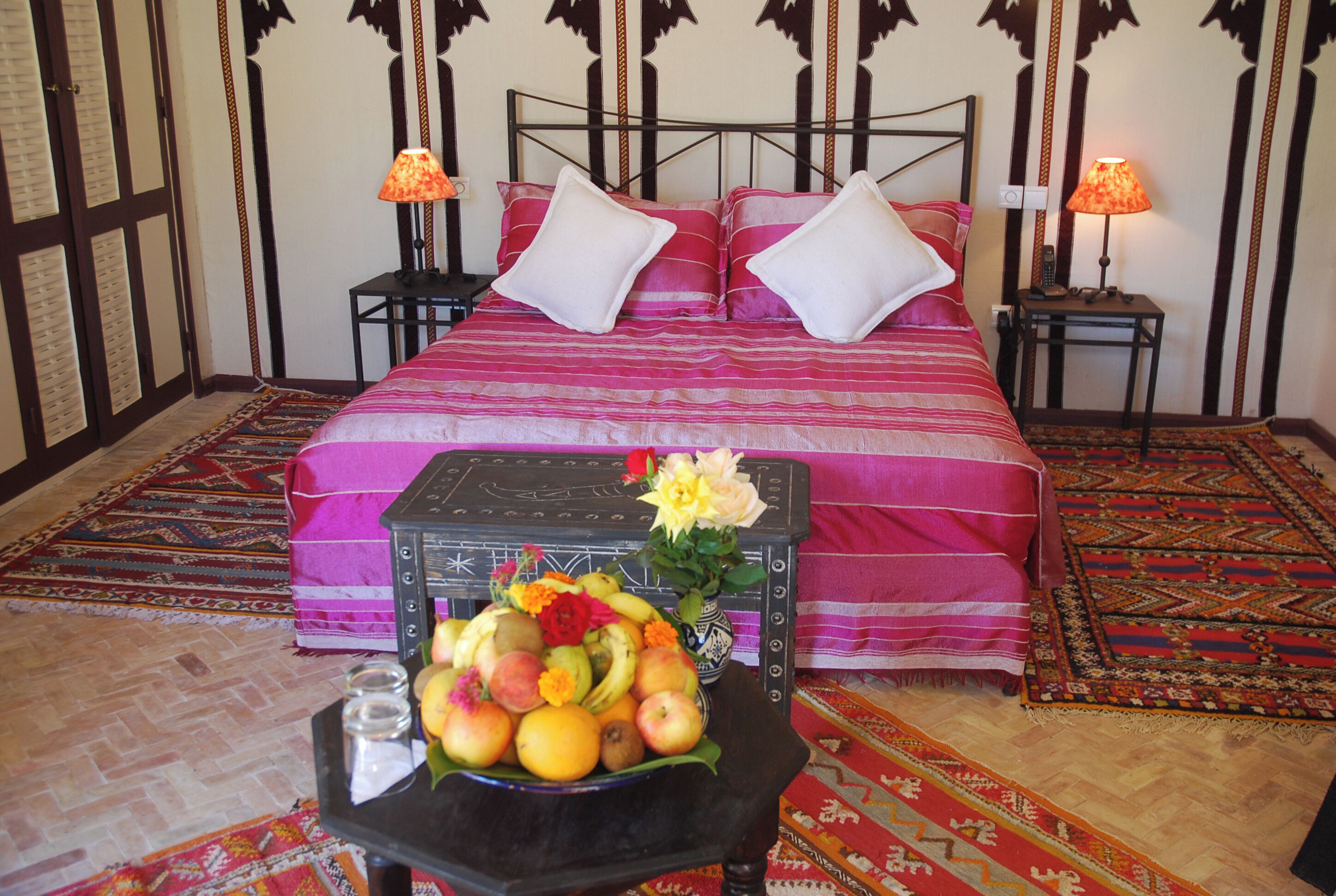 A neatly made bed with a pink bedspread and white pillows, a small table with a fruit bowl and a vase of flowers, and two bedside lamps on nightstands.