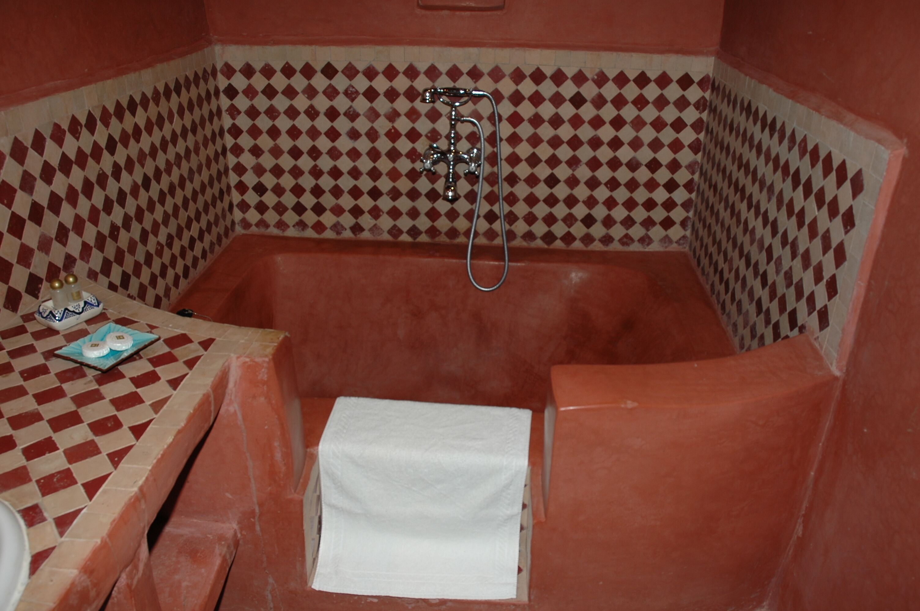 A bathroom with a checkered tile wall, a bathtub, a shower, and a small table with a towel and a bottle.