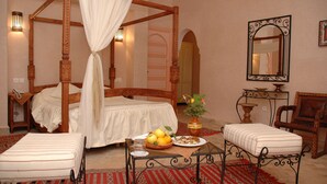 A four-poster bed with a canopy, a bedside table with a lamp, a small table with a bowl of fruit, and a chair with a cushion.