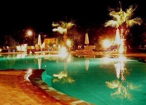Un espace piscine nocturne avec une eau cristalline, des palmiers et des chaises longues.