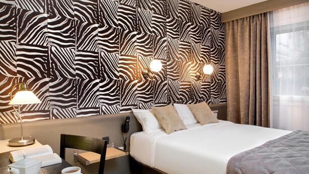 A hotel room with a zebra-patterned wall, a bed with white bedding, a bedside table with a lamp, a chair, a phone, a cup of coffee, and a book. - Logis Hôtel du Midi - Saint Etienne Sud (Saint-Etienne)