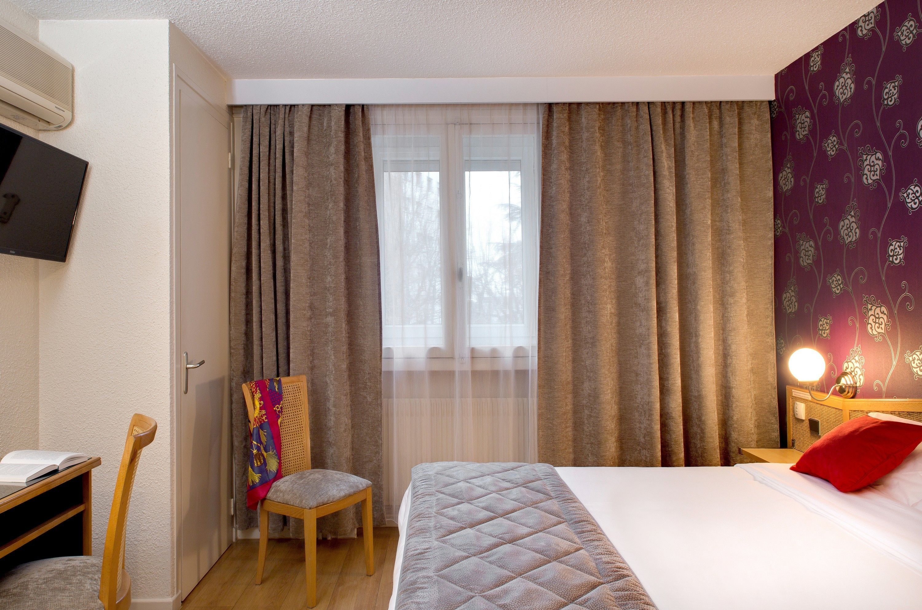 Una habitación de hotel con cama, una silla, un escritorio con un libro, televisión y cortinas.