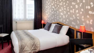 A hotel room with a bed, a chair, a desk with a lamp, and a bookshelf.