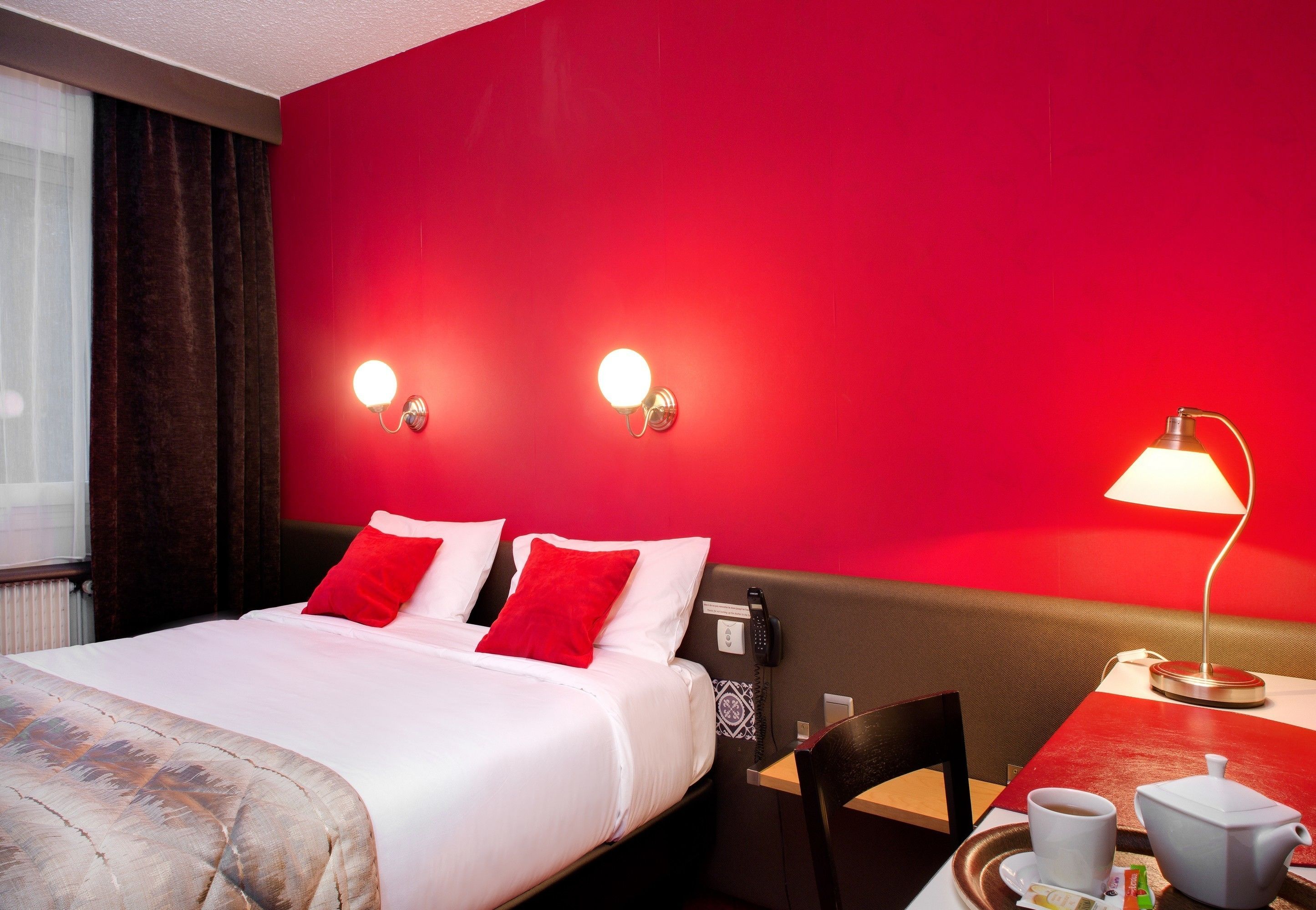 Une chambre d’hôtel avec un mur d’accent rouge, un lit avec une literie blanche et des oreillers rouges, un bureau avec une lampe, une chaise et une table avec une tasse et une théière.