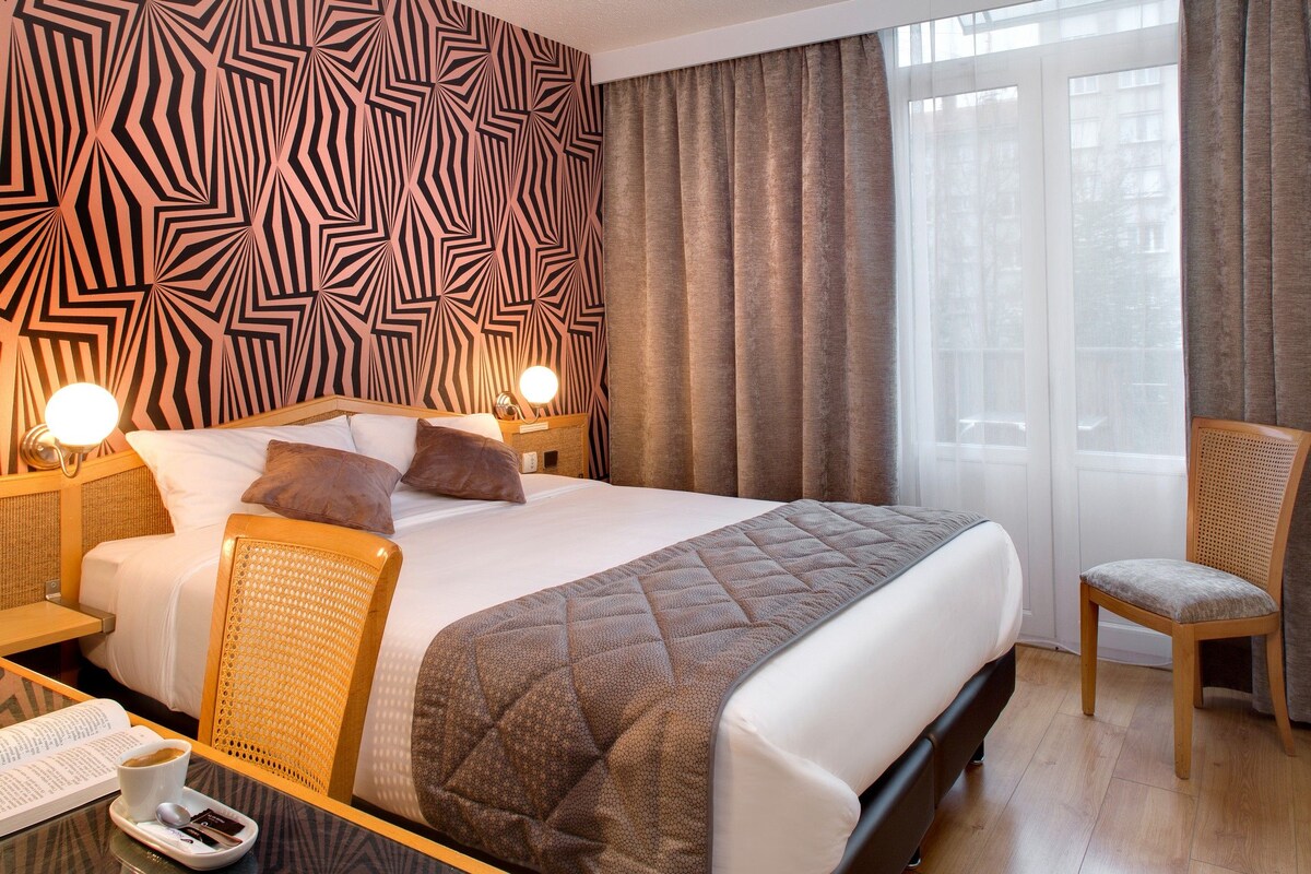 Una habitación de hotel con una cama grande, dos mesitas de noche con lámparas, una silla y vistas al exterior a través de una ventana.