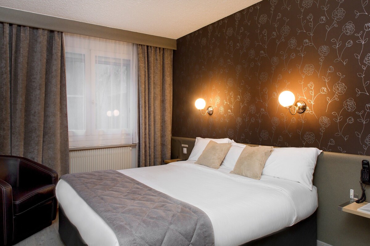 Habitación de hotel con cama, dos lámparas de noche, mesita de noche, una silla y una ventana con cortinas.