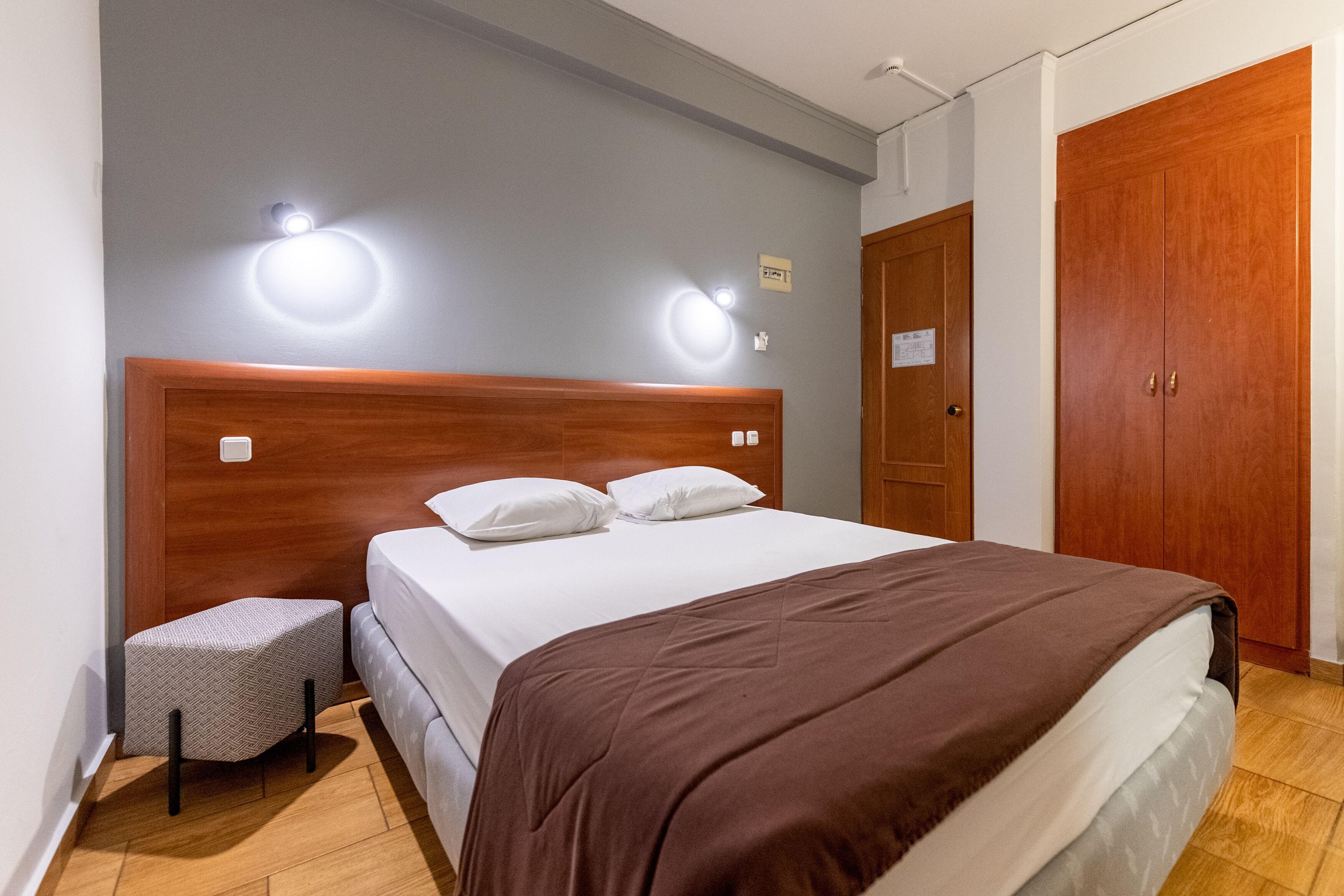 Ein Hotelzimmer mit einem Holzkopfteil, einem Bett mit weißer Bettwäsche, einer braunen Tagesdecke, einem kleinen Nachttisch mit einem gemusterten Kissen und einem Holzschrank.