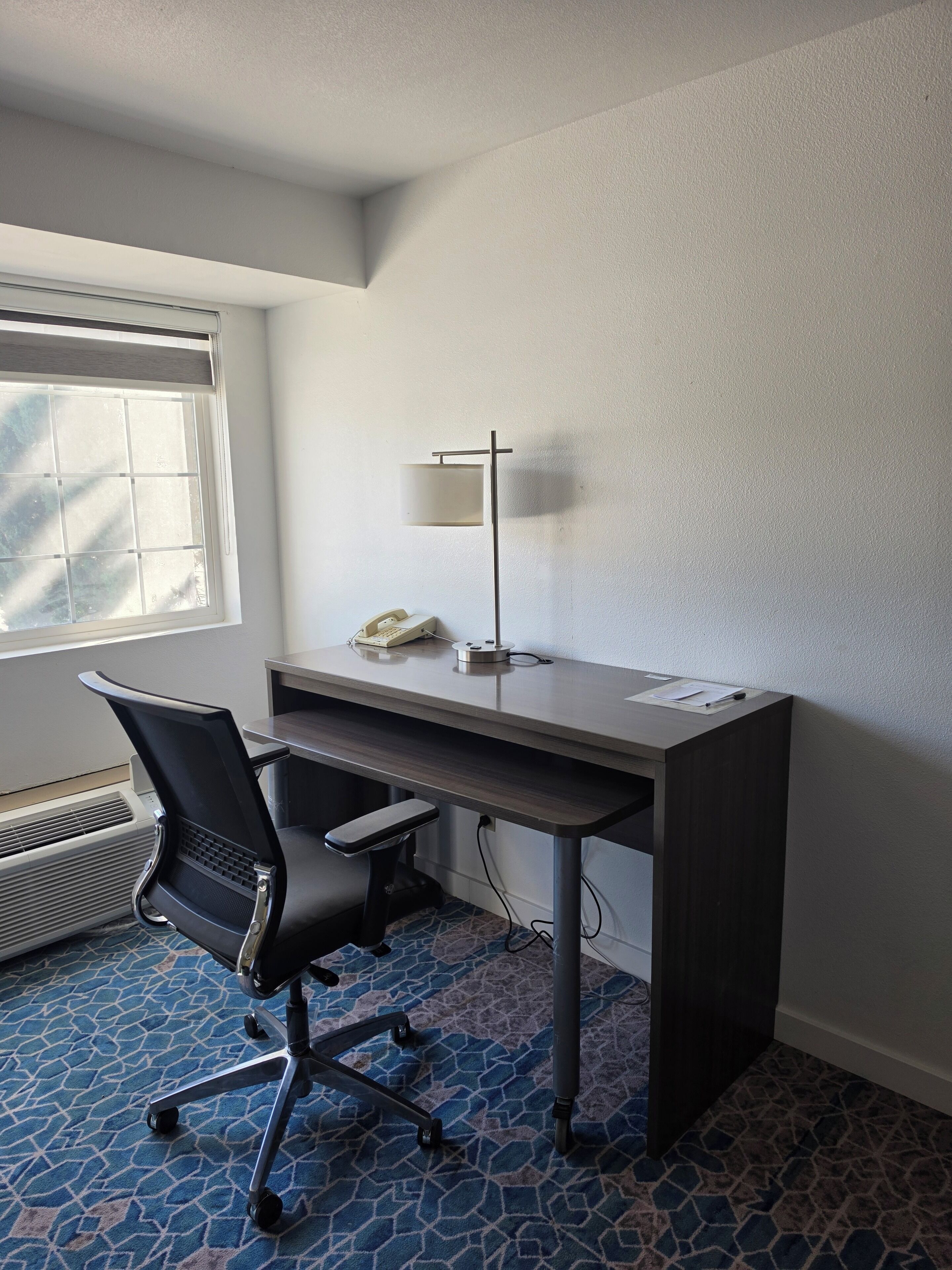 A hotel room with a desk, chair, lamp, phone, and air conditioning unit.