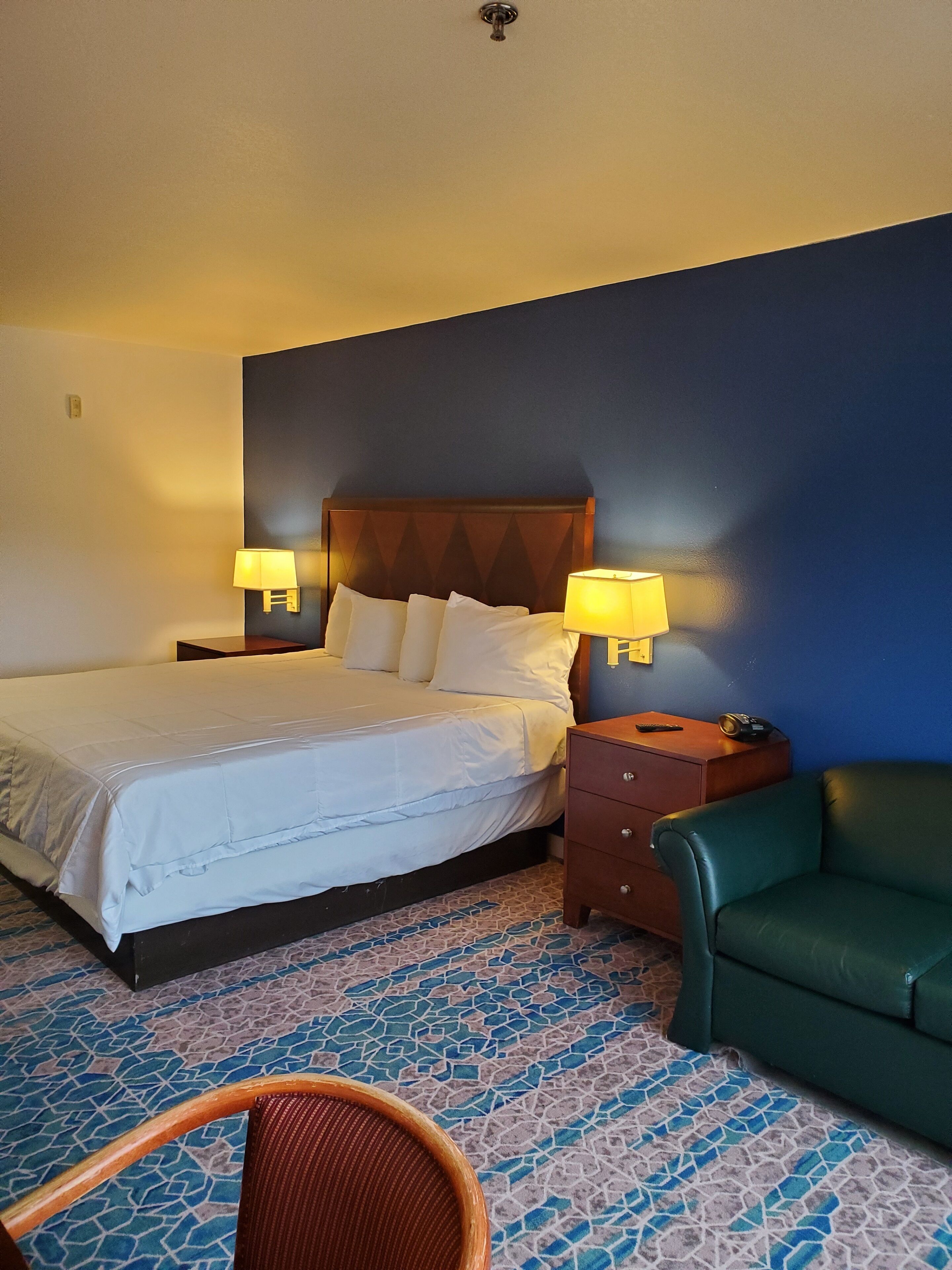 A hotel room with a bed, two bedside lamps, a green chair, and a nightstand with a telephone.
