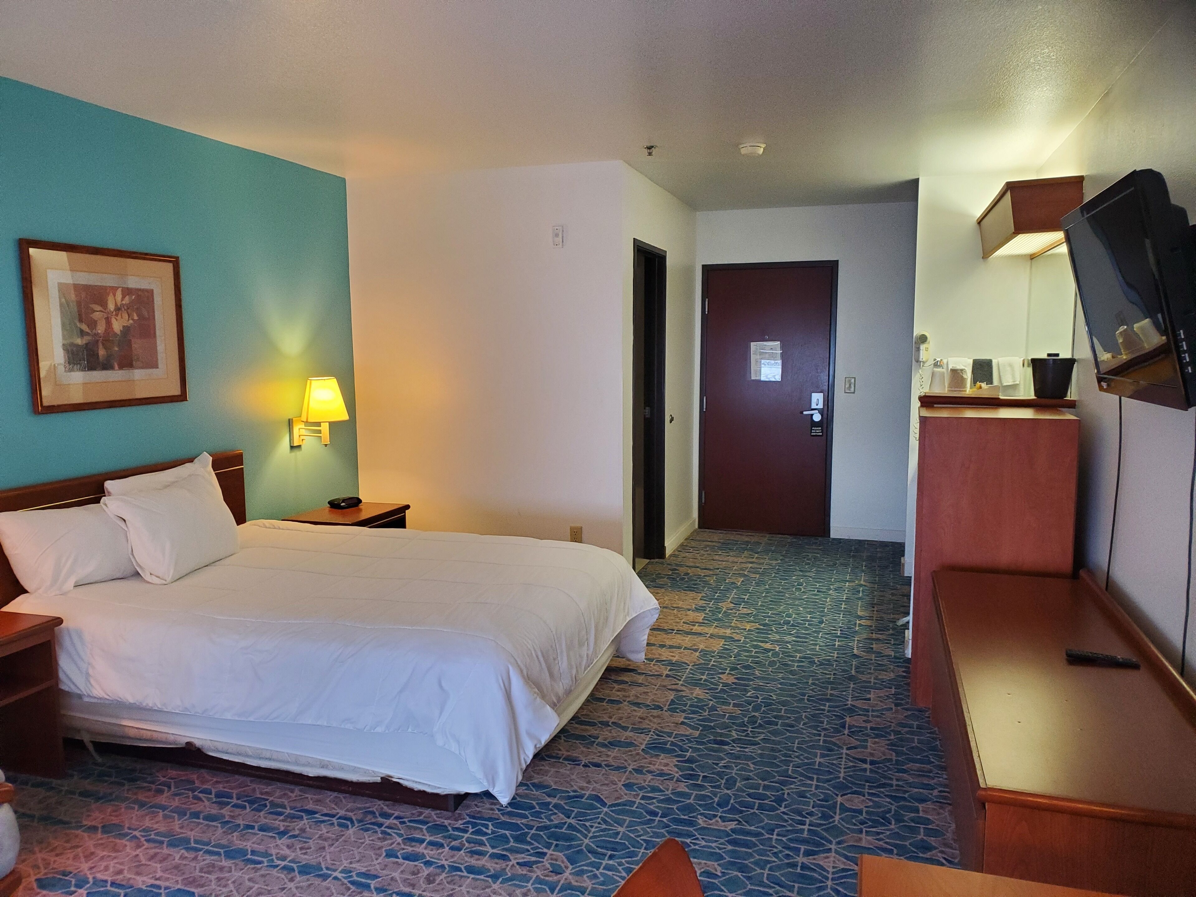 A hotel room with a bed, a nightstand, a lamp, a television, and a door leading to another room.