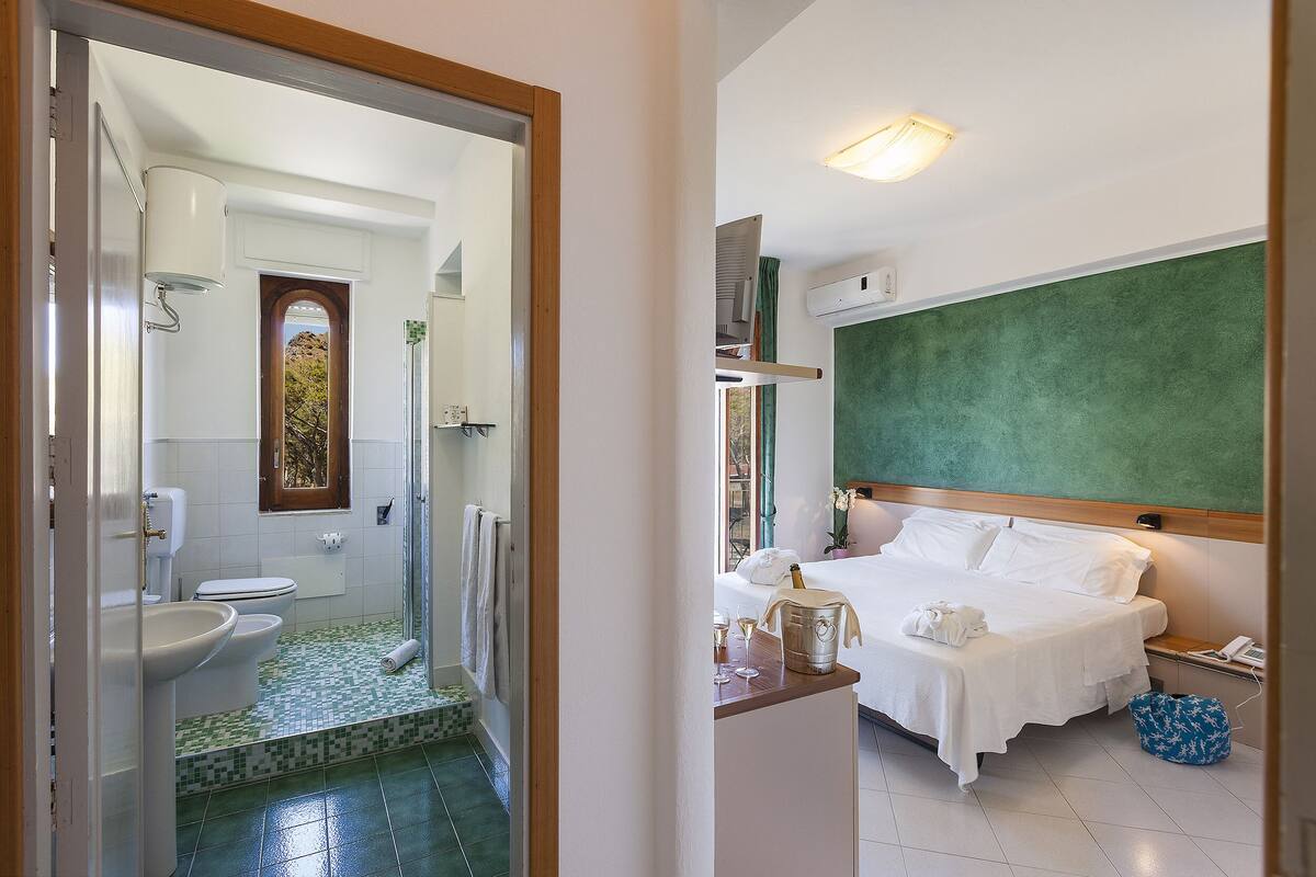 A hotel room with a bed, a bedside table with a bottle and glasses, a bathroom with a toilet, sink, and shower.