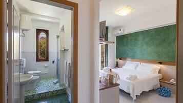 A hotel room with a bed, a bedside table with a bottle and glasses, a bathroom with a toilet, sink, and shower.