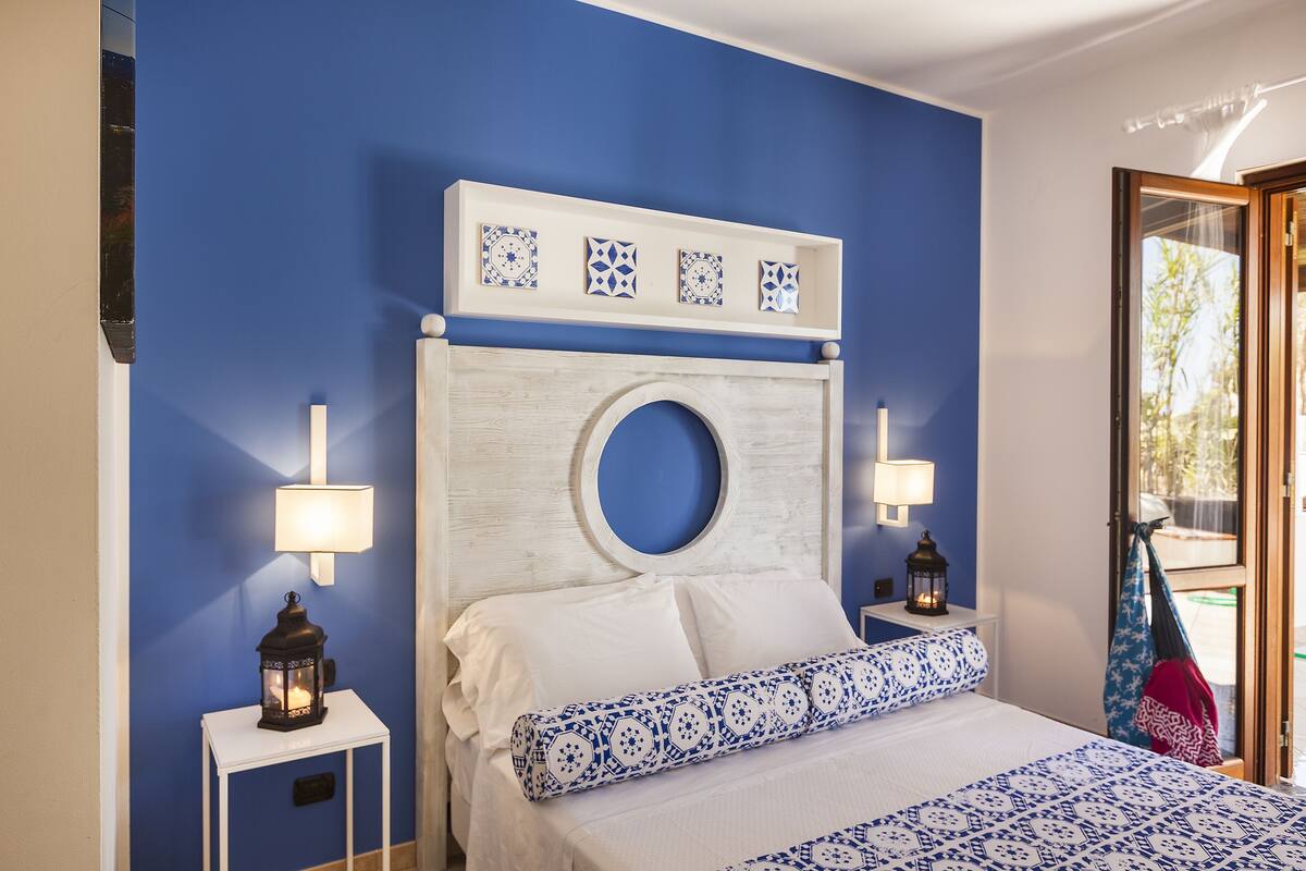 A bedroom with a blue wall, a circular headboard, and a bed with blue and white bedding. There are two bedside tables with lamps and a hanging chair visible.