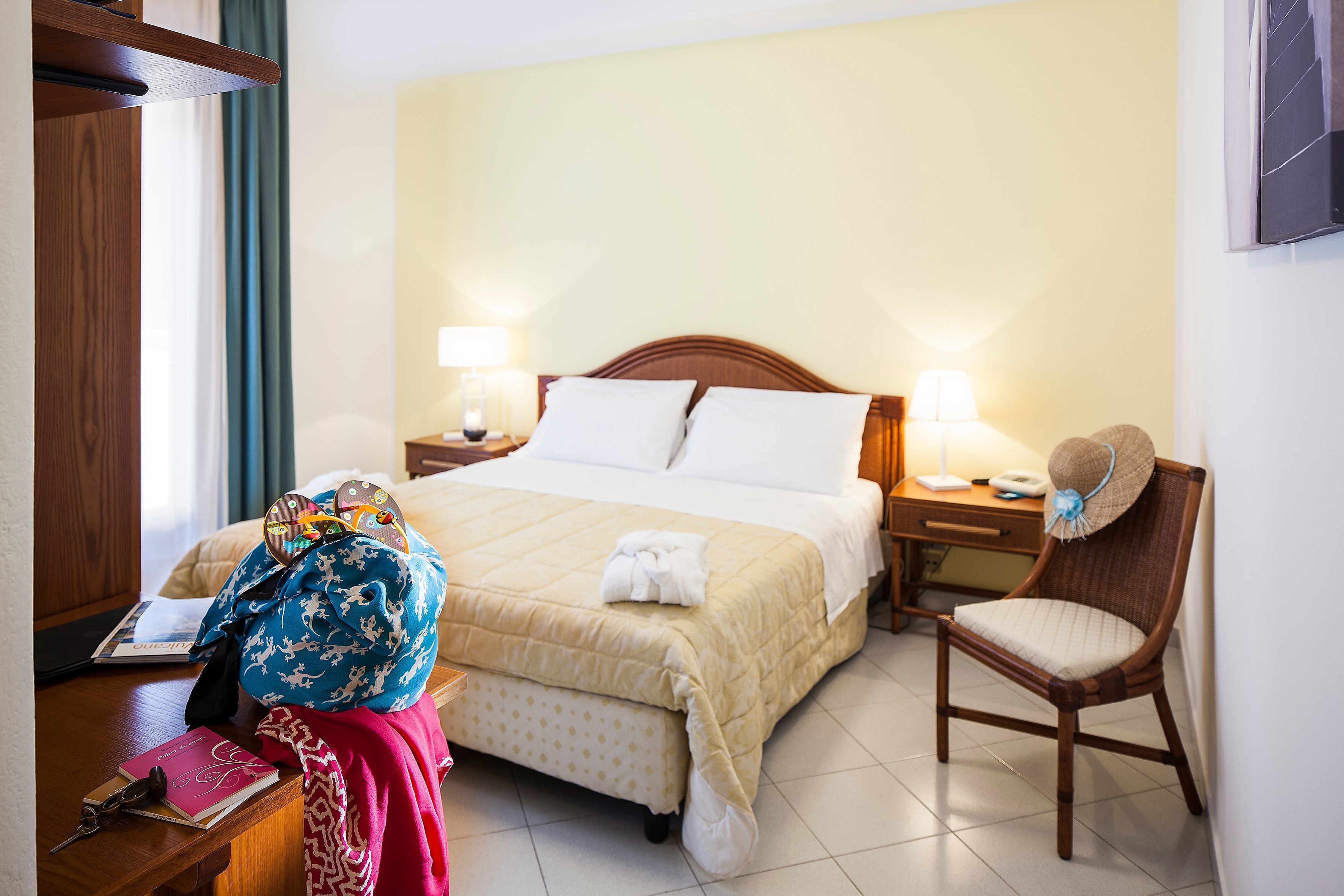 A hotel room with a bed, two bedside lamps, a chair, a desk with a laptop, and a window with curtains.