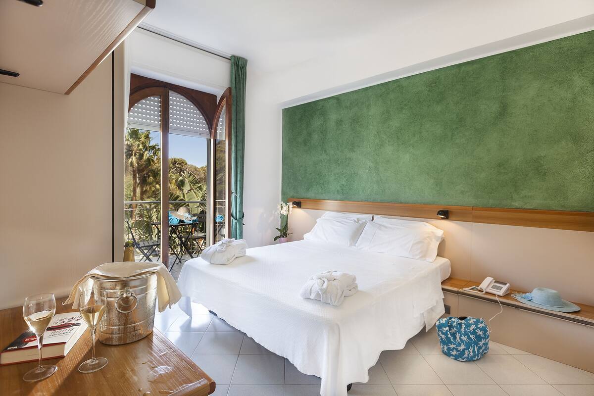A neatly made bed with white linens, a bedside table with a phone and a hat, and a view of the outdoors through a glass door.