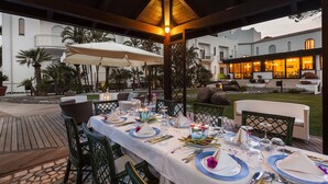 Un área de comedor al aire libre con una sección cubierta, mesas dispuestas con manteles blancos y vistas a un edificio en el fondo.