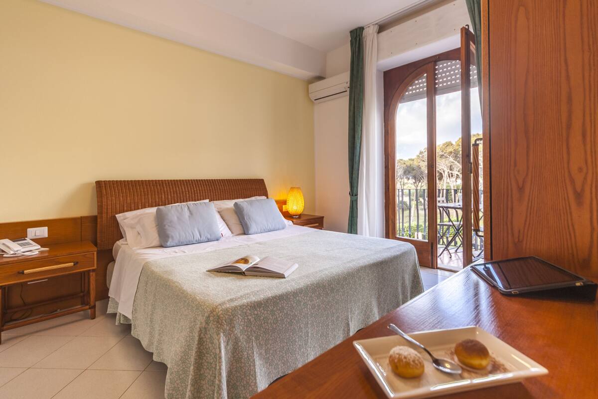 A hotel room with a bed, bedside table, lamp, and a view of the outdoors.