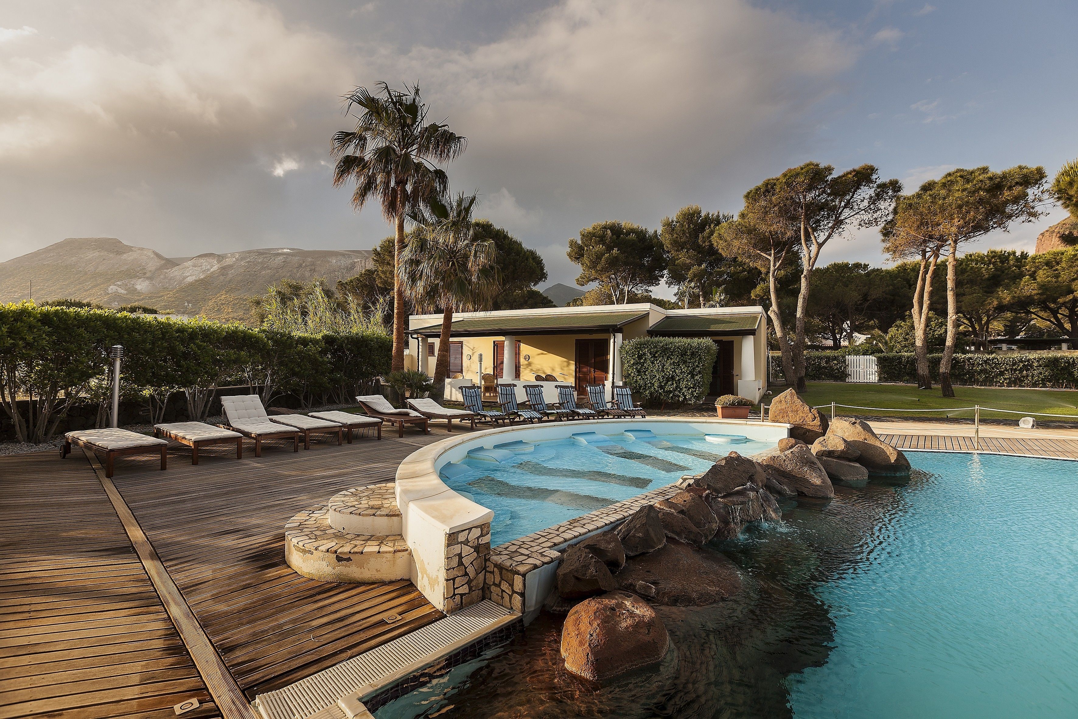 A pool area with lounge chairs, a stone deck, and a building with a view of mountains.
