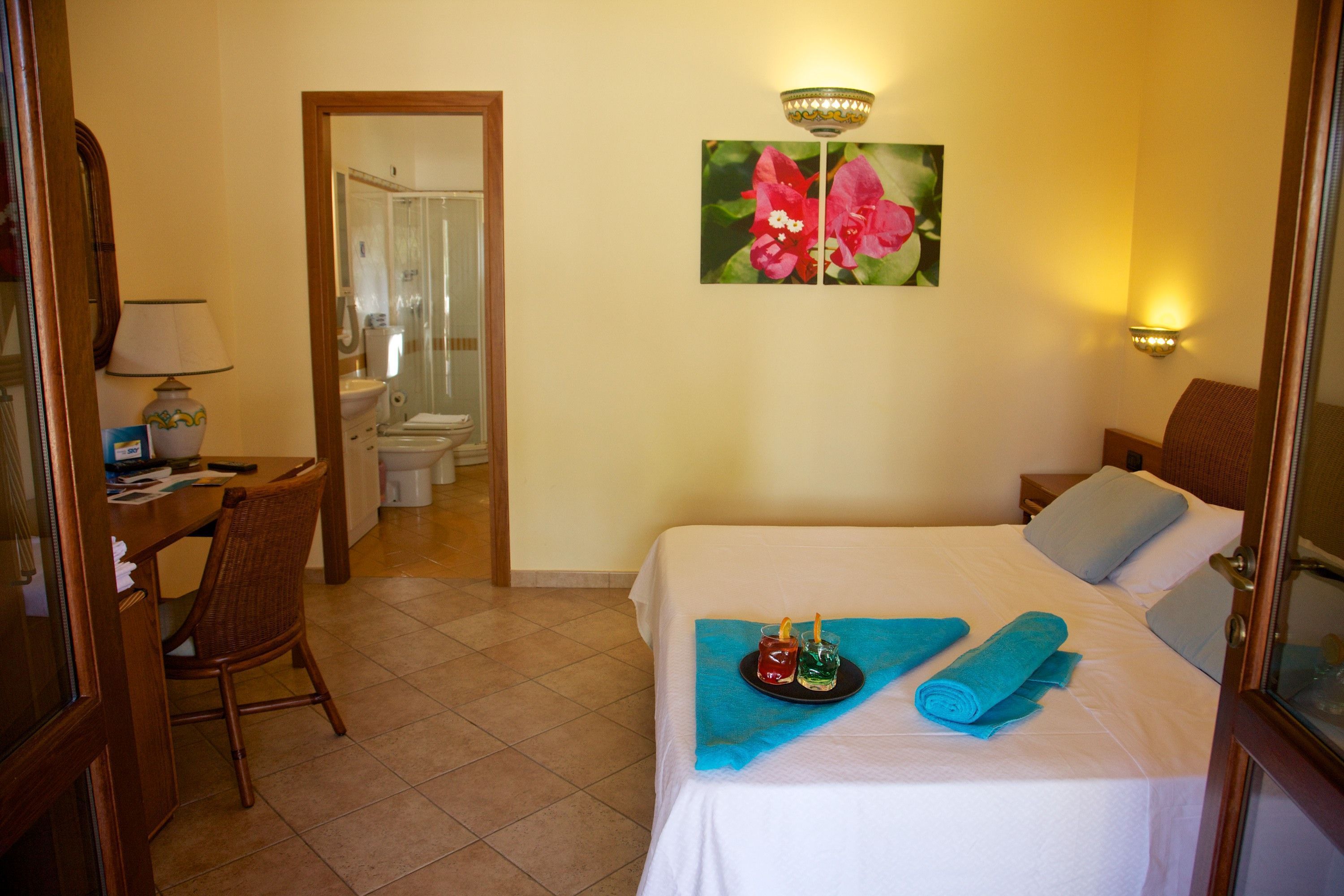 Una habitación de hotel con cama, escritorio, silla, baño visible a través de una puerta abierta y un cuadro de flores en la pared.