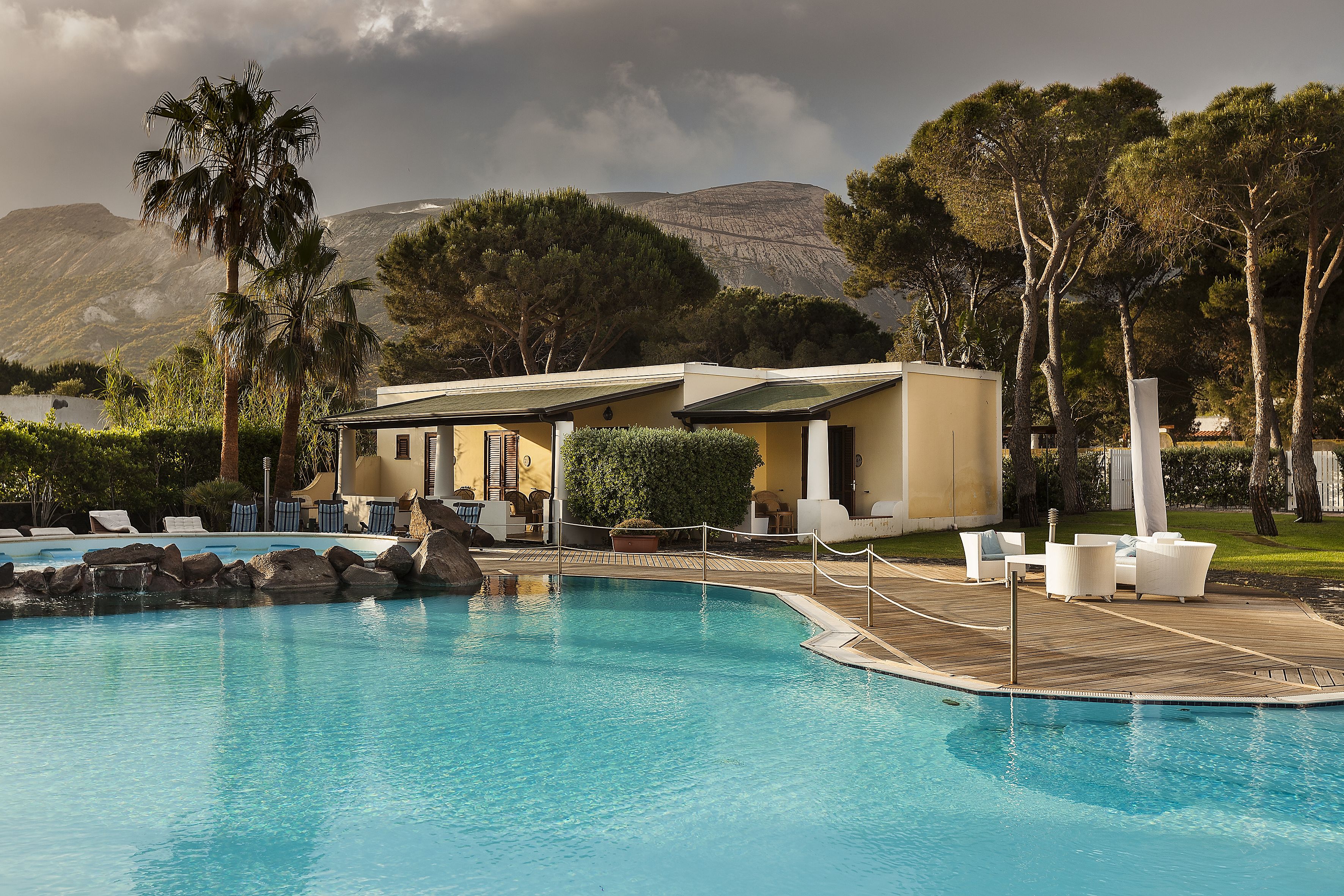 A pool area with lounge chairs, a small building, and a mountainous backdrop.