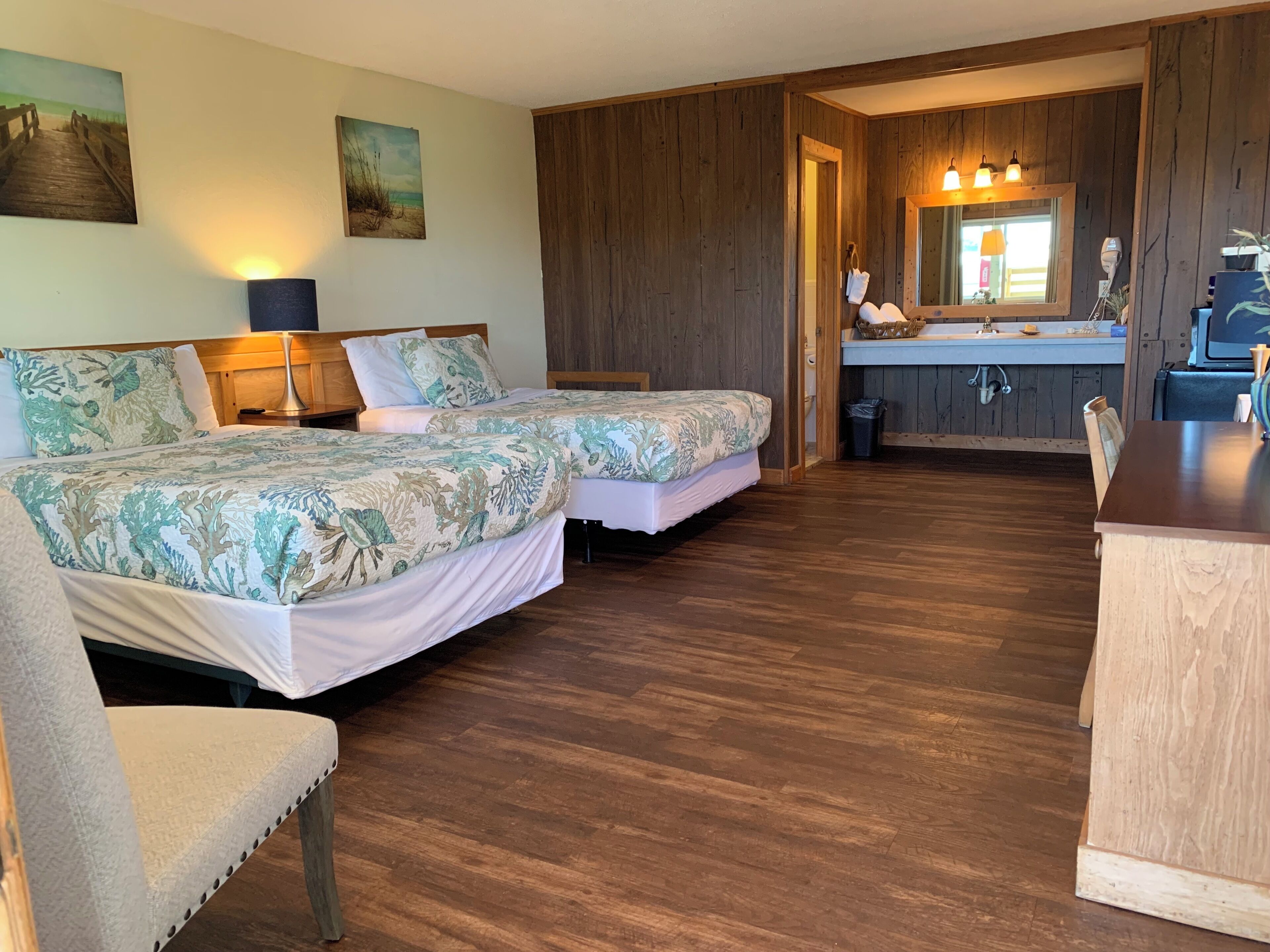 Una habitación con dos camas, suelo de madera, un escritorio pequeño, un espejo y un lavabo.