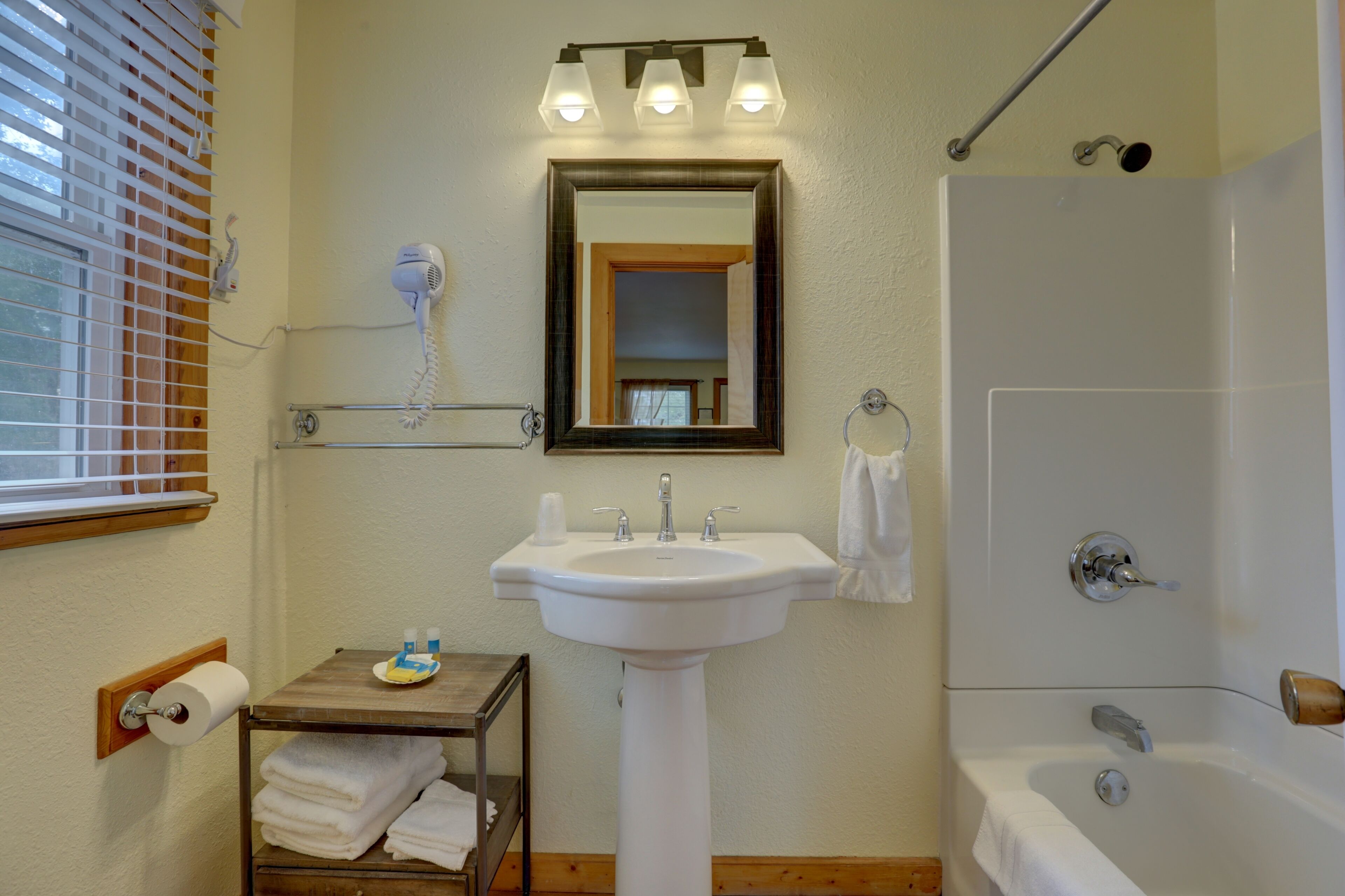 A bathroom with a sink, mirror, and shower. There is a towel rack, a hairdryer, and a small table with towels.