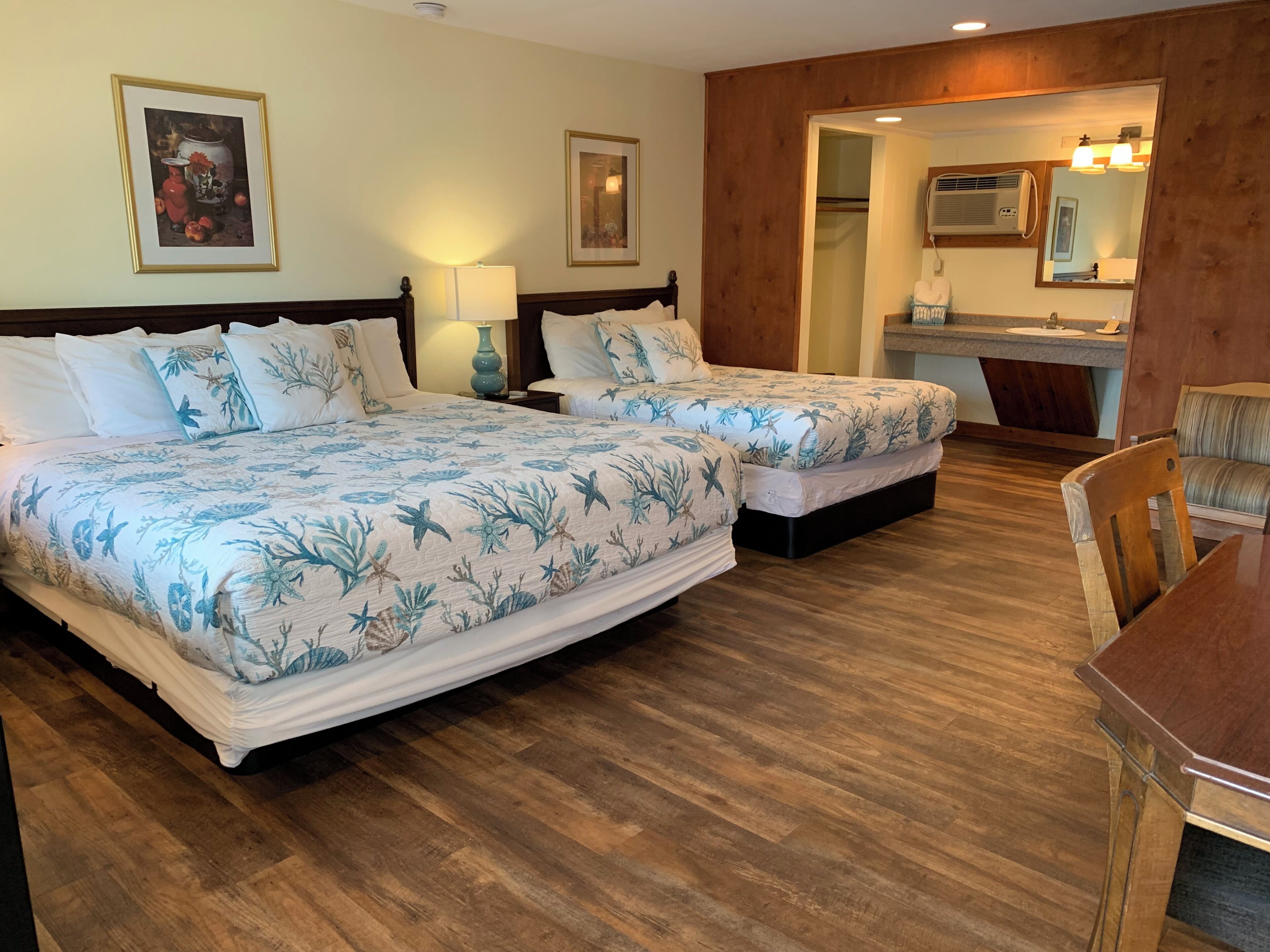 A hotel room with two beds, a wooden floor, a desk, and a bathroom visible through an open door.