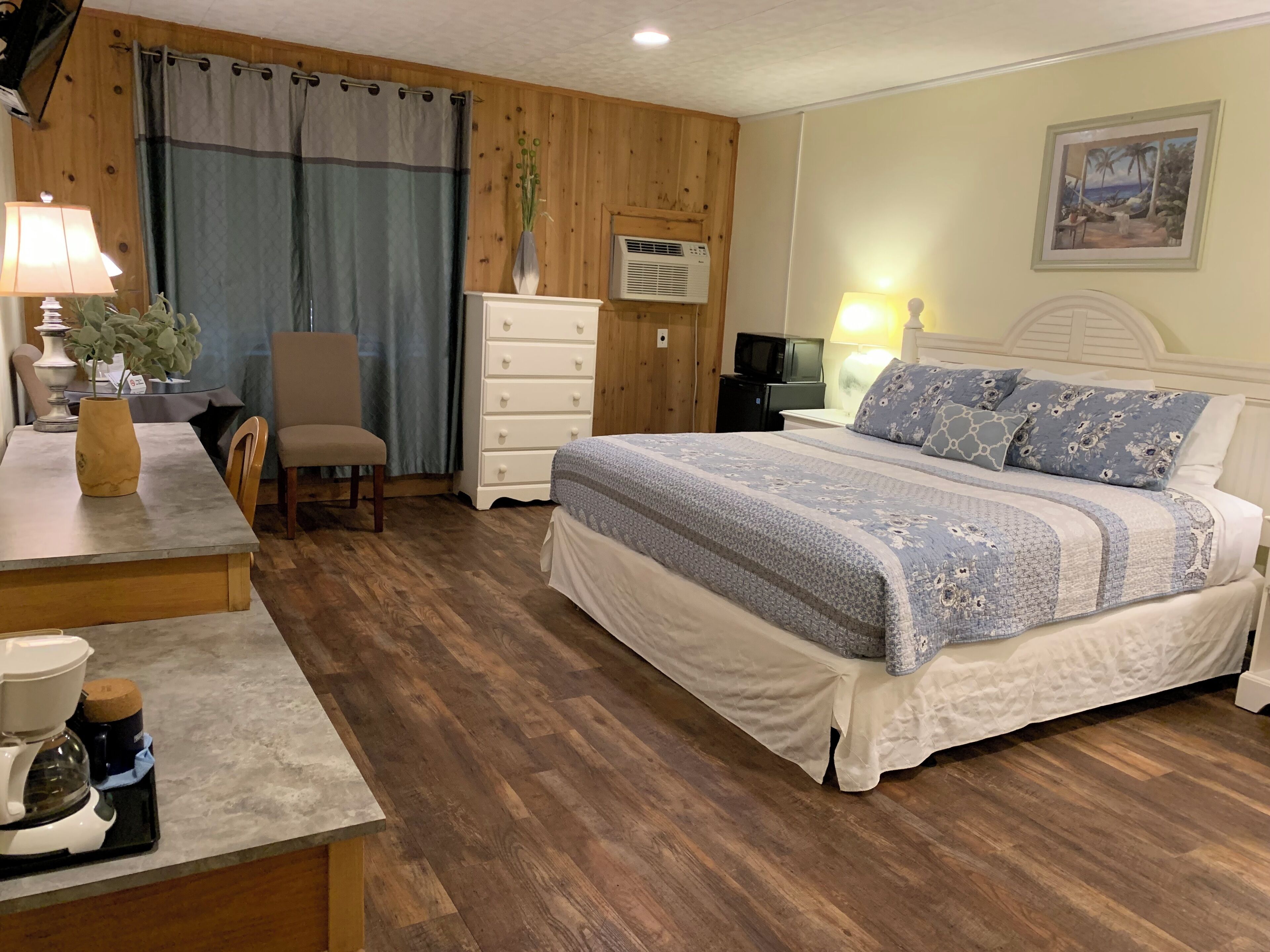 A bedroom with a bed, dresser, chair, desk, coffee maker, and a painting on the wall.