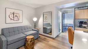 A modern living room with a grey sofa, a round wooden stool, and a coffee table. There is a mirror reflecting a grey sofa and a small table. The room has wooden flooring and a white ceiling. - Country Inn & Suites by Radisson, Savannah Airport, GA (Savannah)