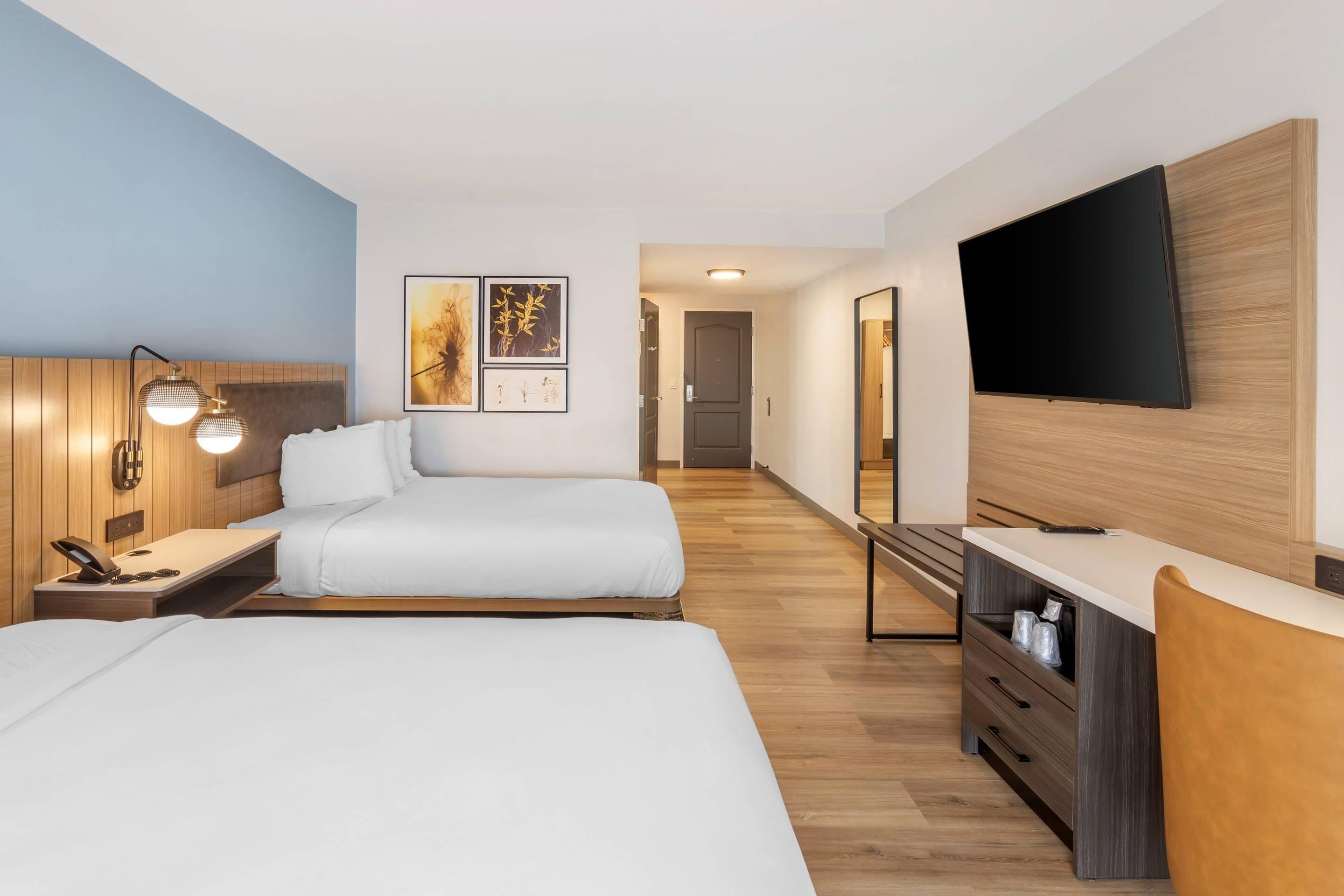 A hotel room with two beds, a desk, a TV, and artwork on the wall.