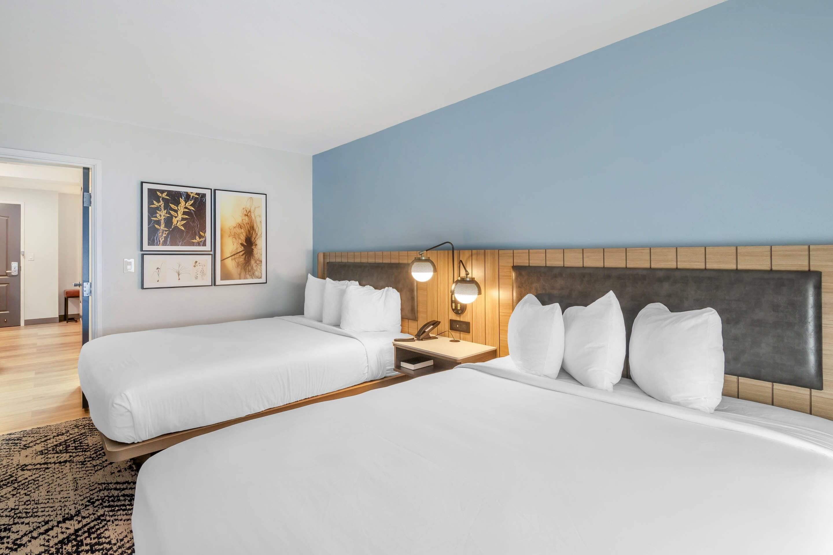 A hotel room with two beds, a wooden headboard, wall-mounted artwork, a bedside table with a lamp, and a patterned rug.
