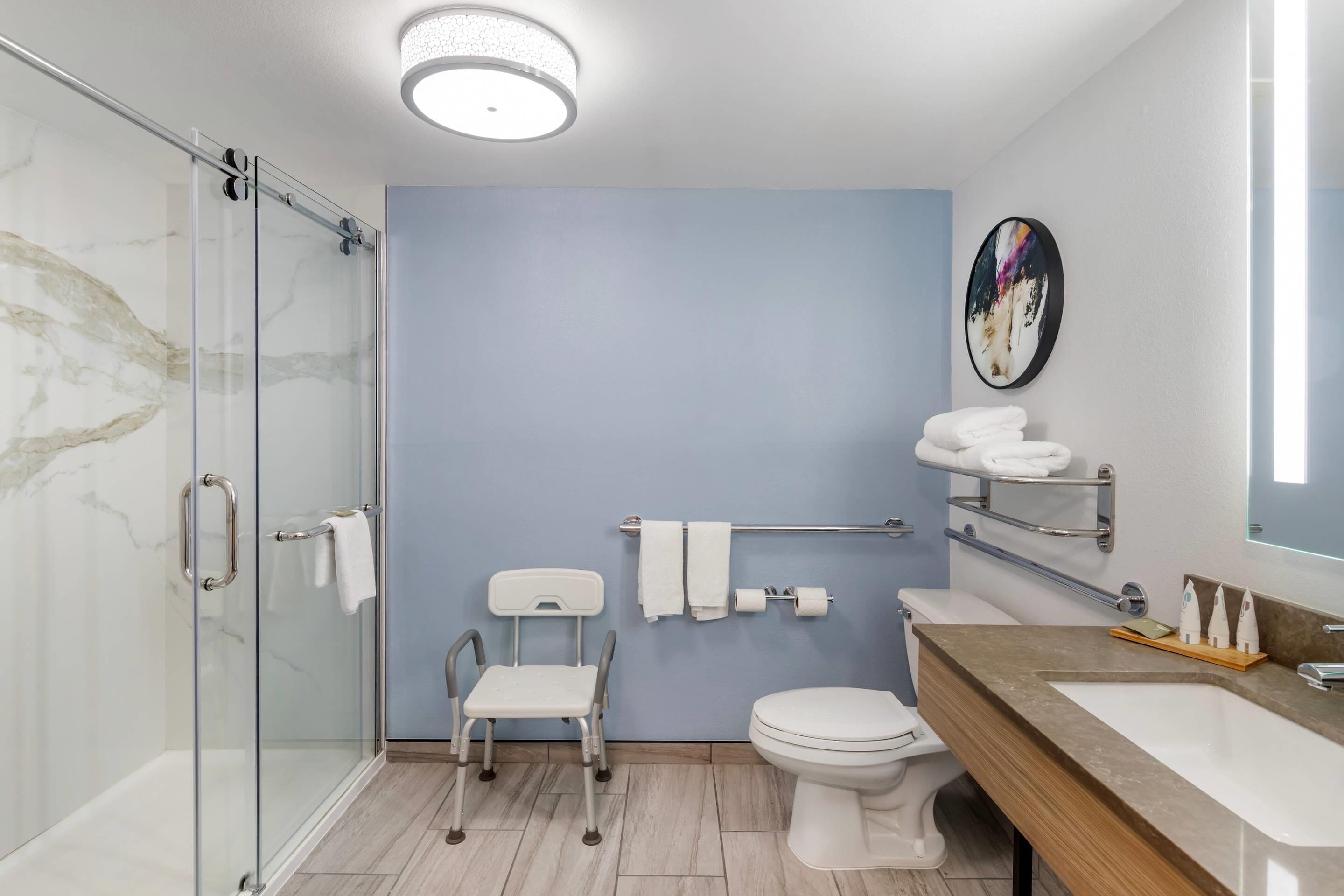 Une salle de bain moderne dotée d’une douche à parois de verre, d’un siège adapté aux personnes en fauteuil roulant, de toilettes et d’un meuble-lavabo avec un miroir.
