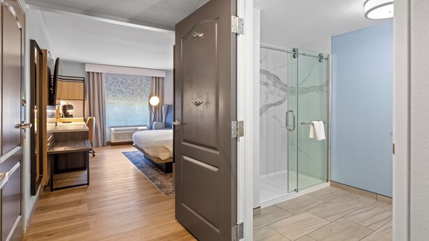 A hotel room with a wooden floor, a bed with a headboard, a desk with a chair, a glass shower enclosure, and a view of the city. - Country Inn & Suites by Radisson, Savannah Airport, GA (Savannah)
