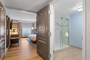 A hotel room with a wooden floor, a bed with a headboard, a desk with a chair, a glass shower enclosure, and a view of the city. - Country Inn & Suites by Radisson, Savannah Airport, GA (Savannah)