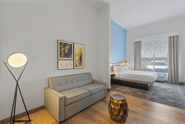 A hotel room with a grey sofa, a round floor lamp, a decorative stool, and a bed with a view of the outside.