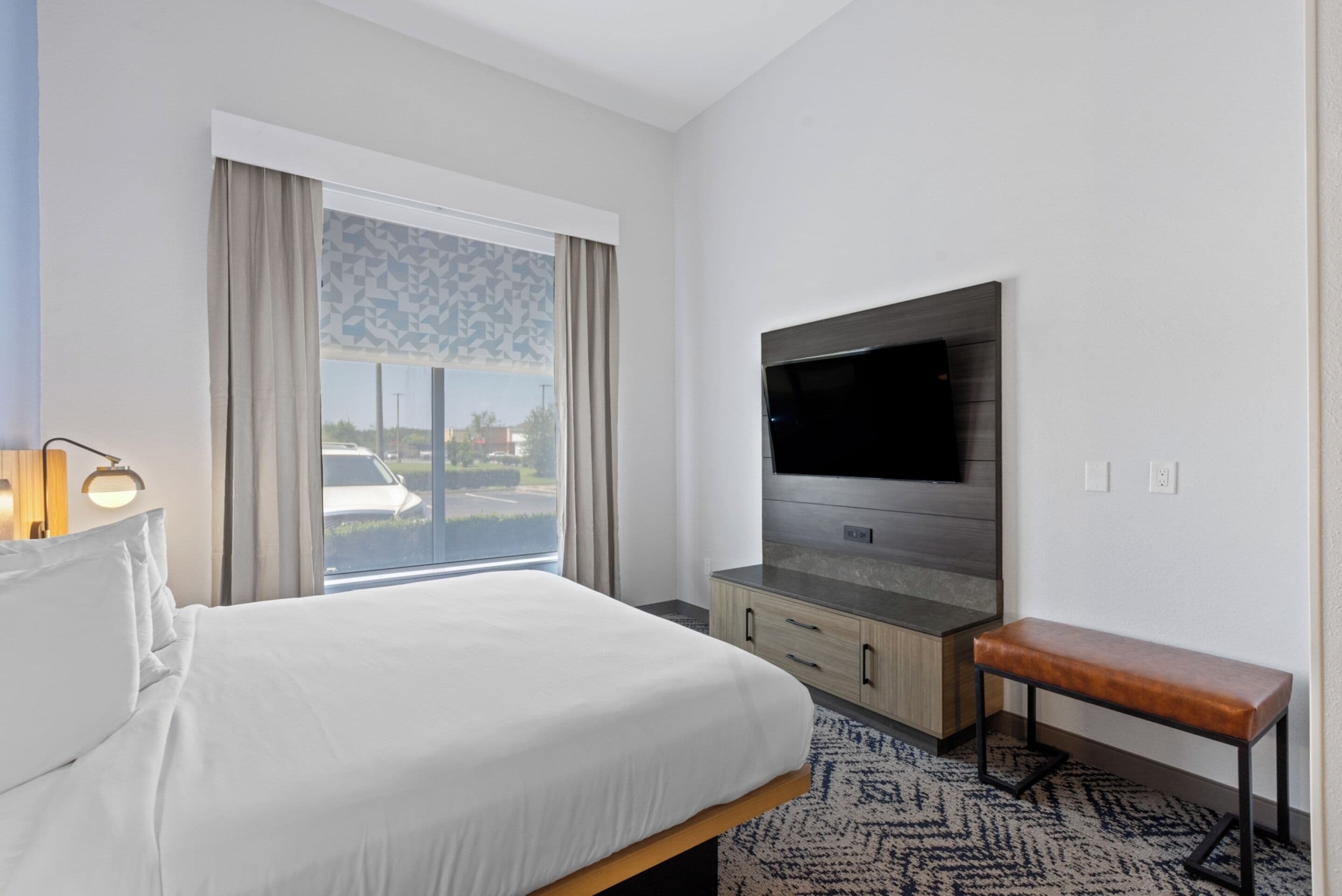 A hotel room with a bed, a TV, a bench, and a window with curtains.