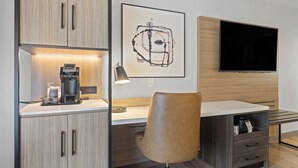A modern hotel room with a wooden cabinet, a coffee machine, a desk with a chair, a framed abstract artwork, and a flat-screen TV mounted on the wall. - Country Inn & Suites by Radisson, Savannah Airport, GA (Savannah)