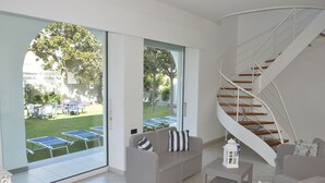 Ein modernes Wohnzimmer mit einer geschwungenen Treppe, einem grauen Sofa mit gestreiften Kissen und Blick auf eine Terrasse mit Tischen und Stühlen.