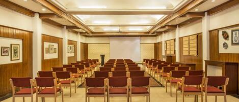 Un salón de conferencias con filas de sillas de madera y una pantalla grande al frente.