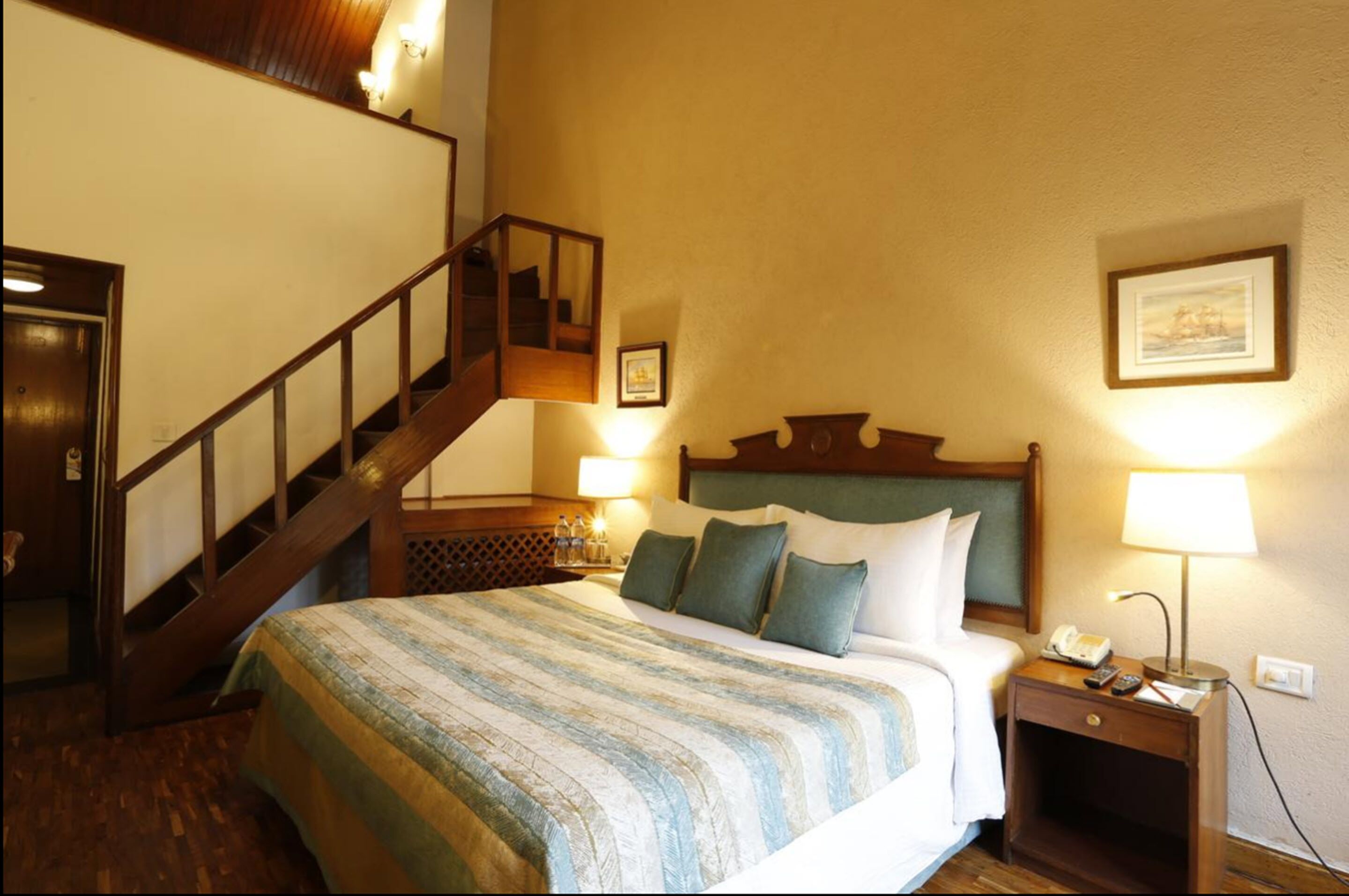 Quarto de hotel com cama, mesa de cabeceira com abajur, escada de madeira e um quadro na parede.