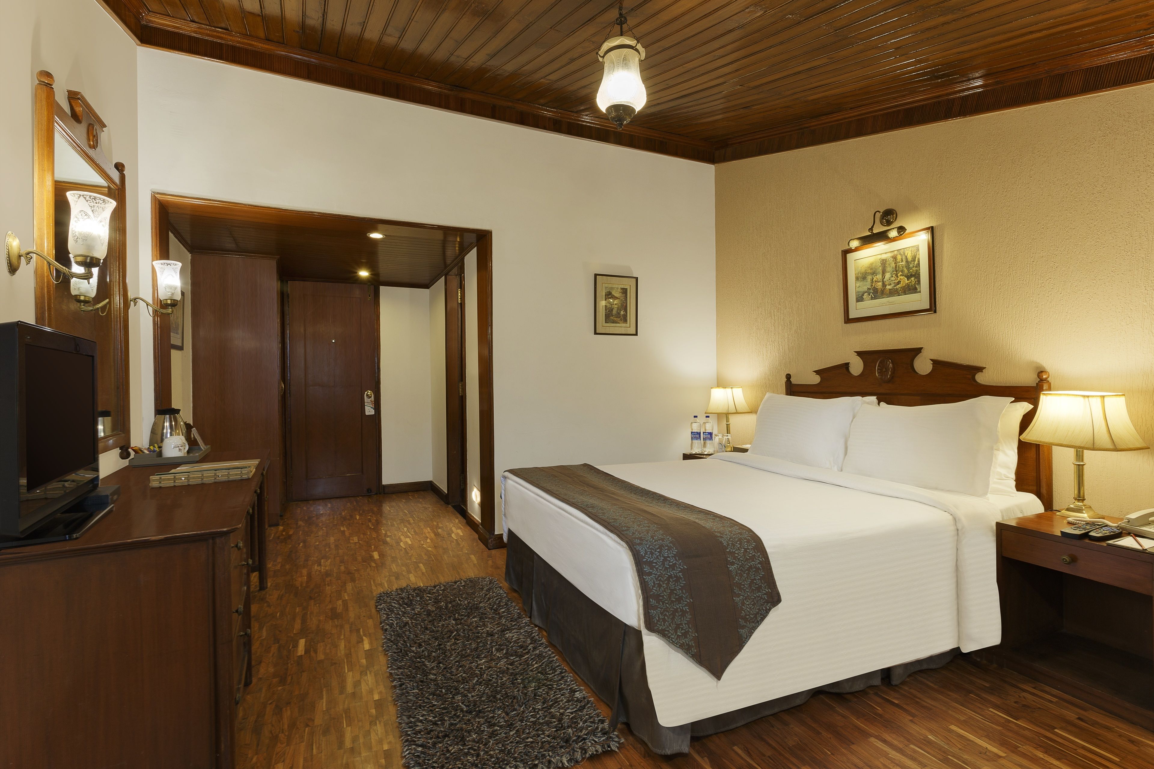 Habitación de hotel con techo de madera, cama con ropa blanca, mesita de noche con lámpara, escritorio con ordenador y una alfombra pequeña.